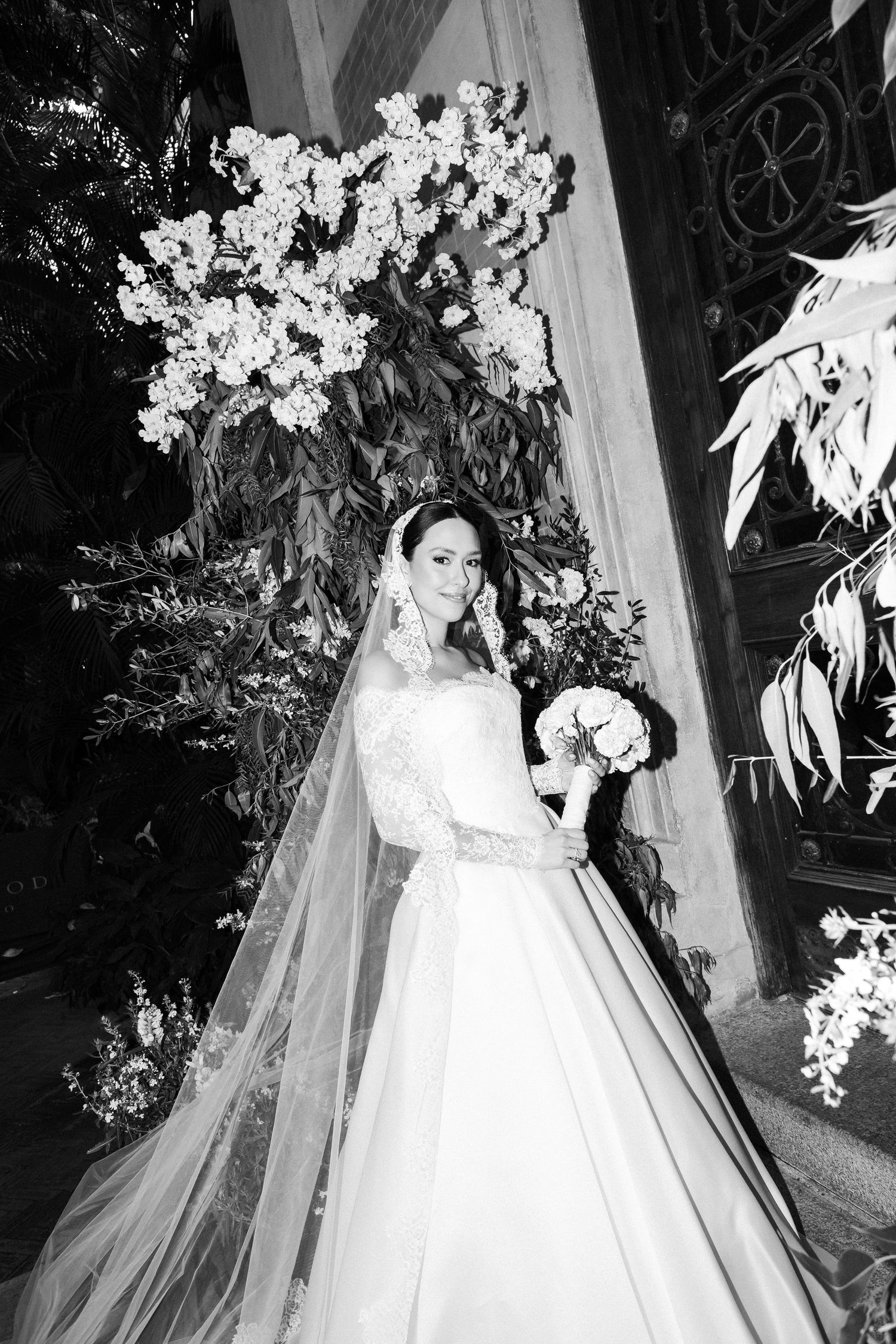 A bride in a classic white ballgown wedding dress and veil poses elegantly against a floral backdrop.