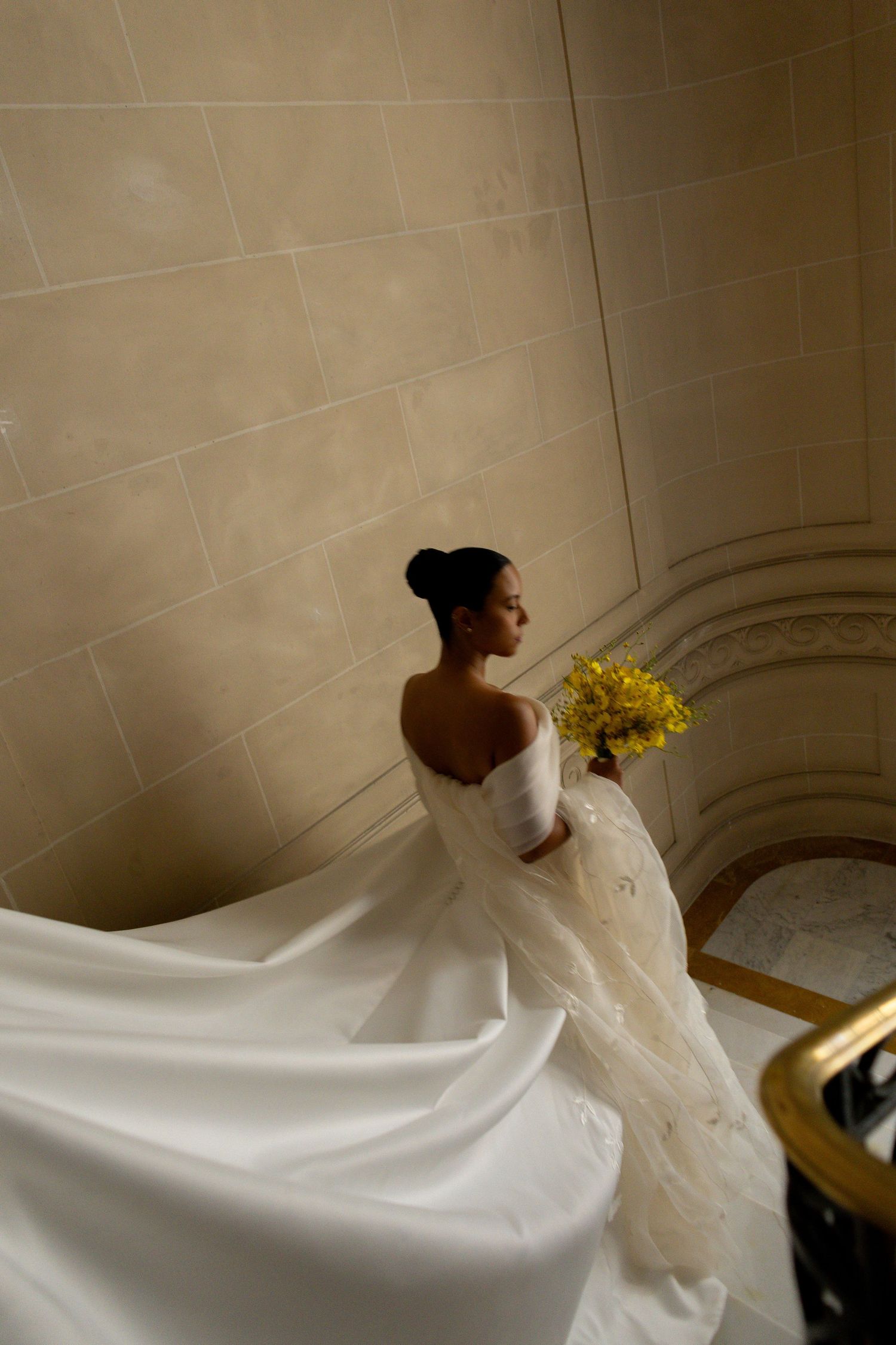 A bride in a flowing white wedding dress holds yellow flowers while ascending elegant stone stairs in dramatic lighting.