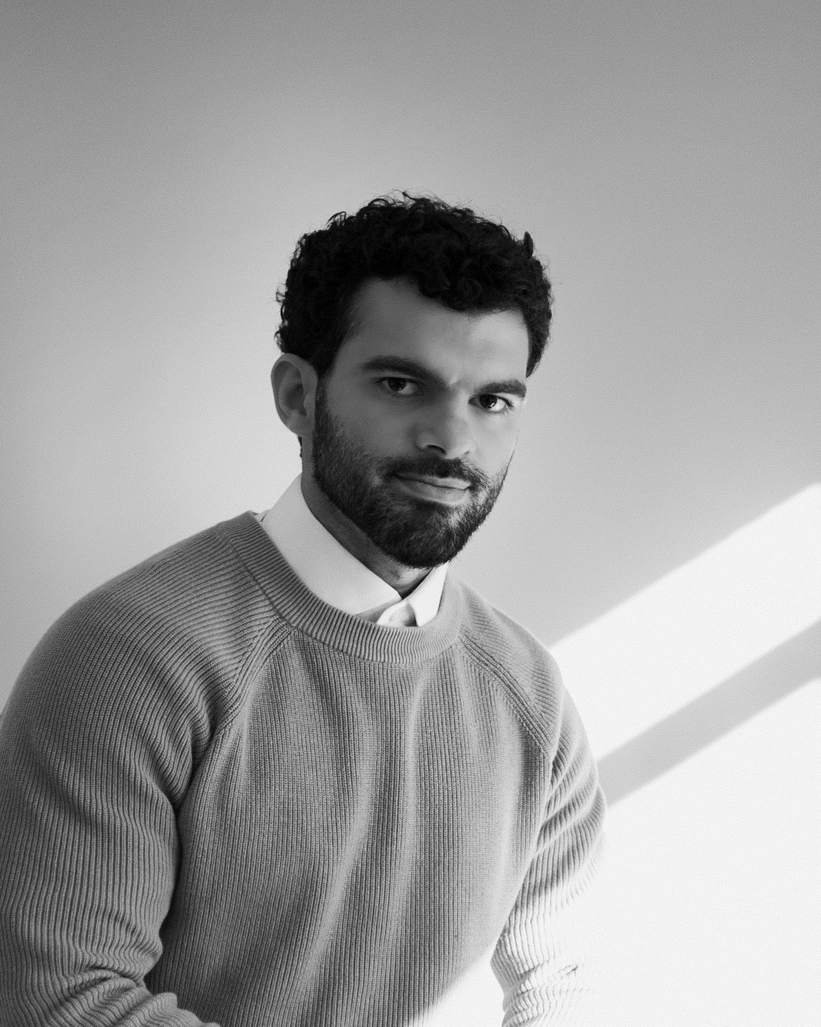 Black and white portrait of a bearded man with curly hair wearing a knit sweater over a collared shirt.