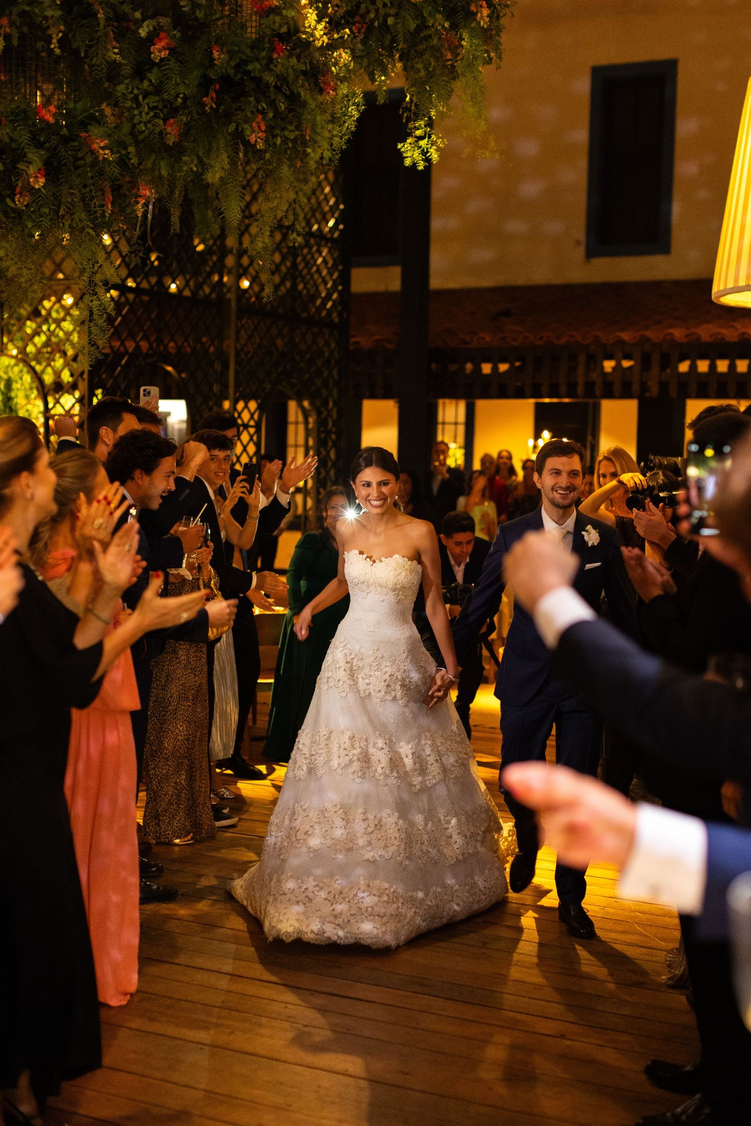 A wedding celebration on a wooden deck at night with guests in formal attire surrounding someone in a lace wedding gown.