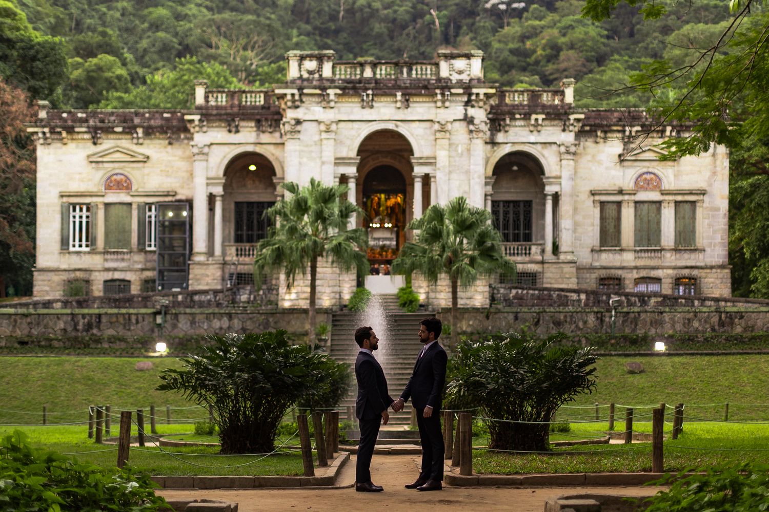 Two people stand in silhouette before an elegant colonial mansion with arched architecture and palm trees in Rio de Janeiro.