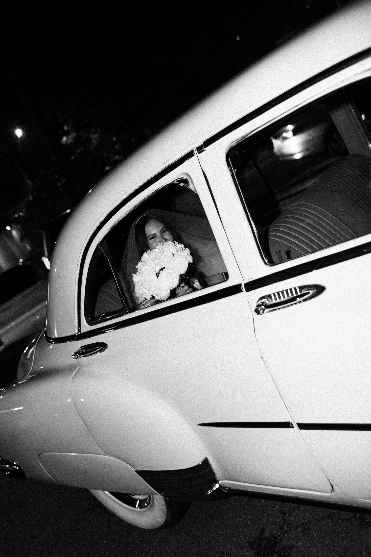 A black and white photo showing a vintage car with a bouquet in the rear window at night.