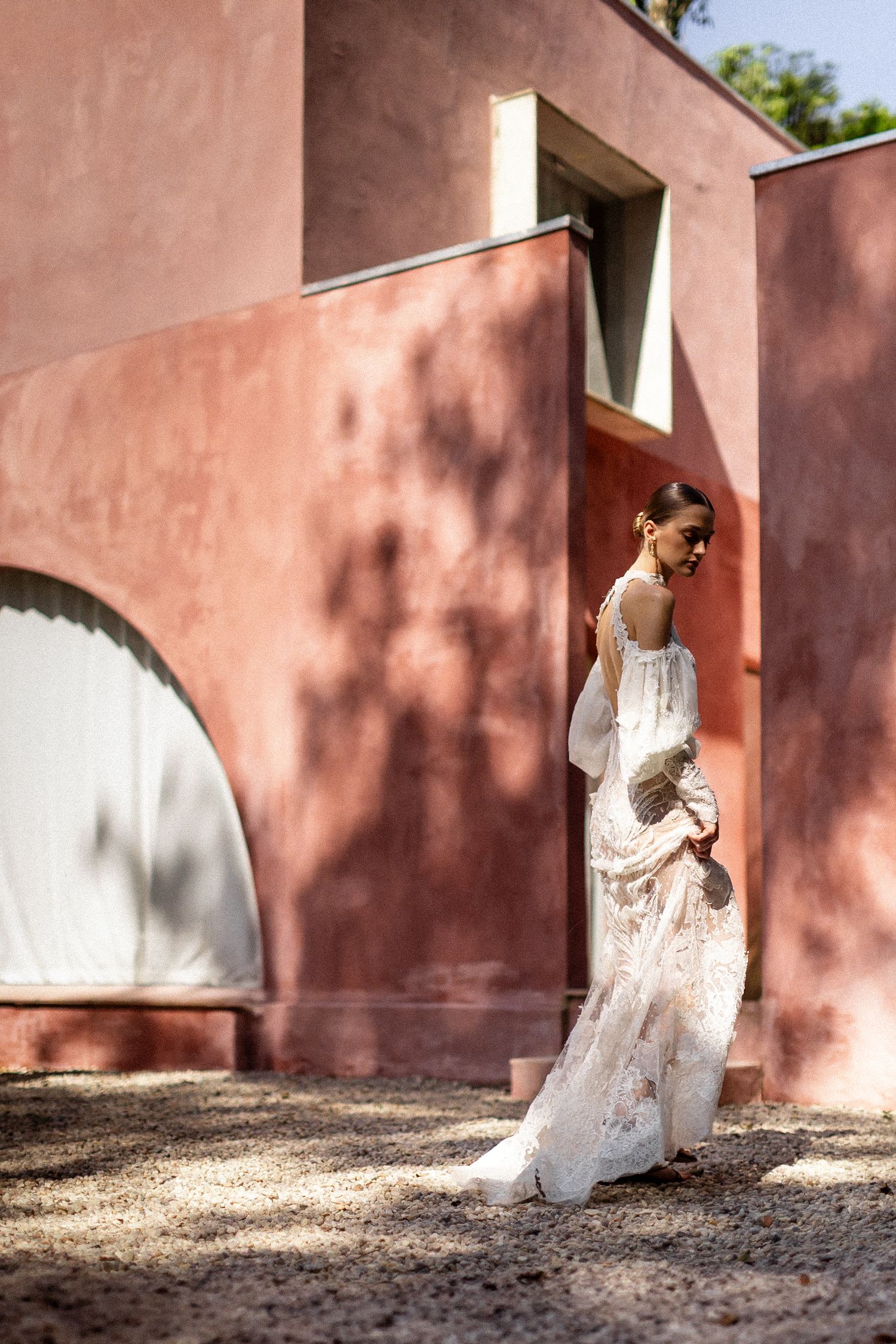 A person in a flowing white lace wedding dress stands against a terracotta-colored wall in dappled sunlight.