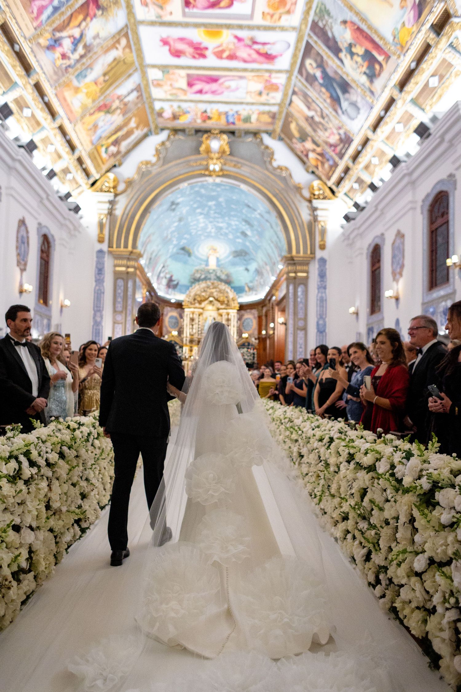 A wedding ceremony in an ornate cathedral with white floral aisle decorations and guests watching from the sides.