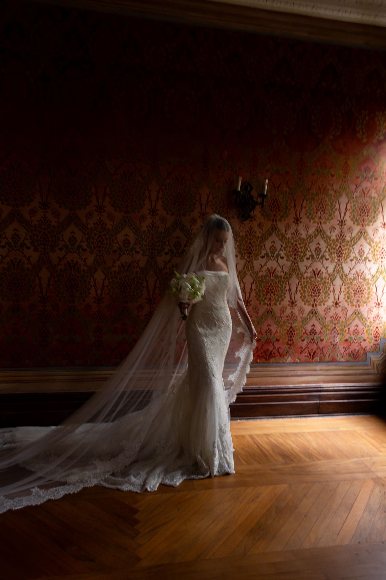 A bride in a fitted lace wedding gown and long flowing veil stands against an ornate damask wallpaper in a historic room.