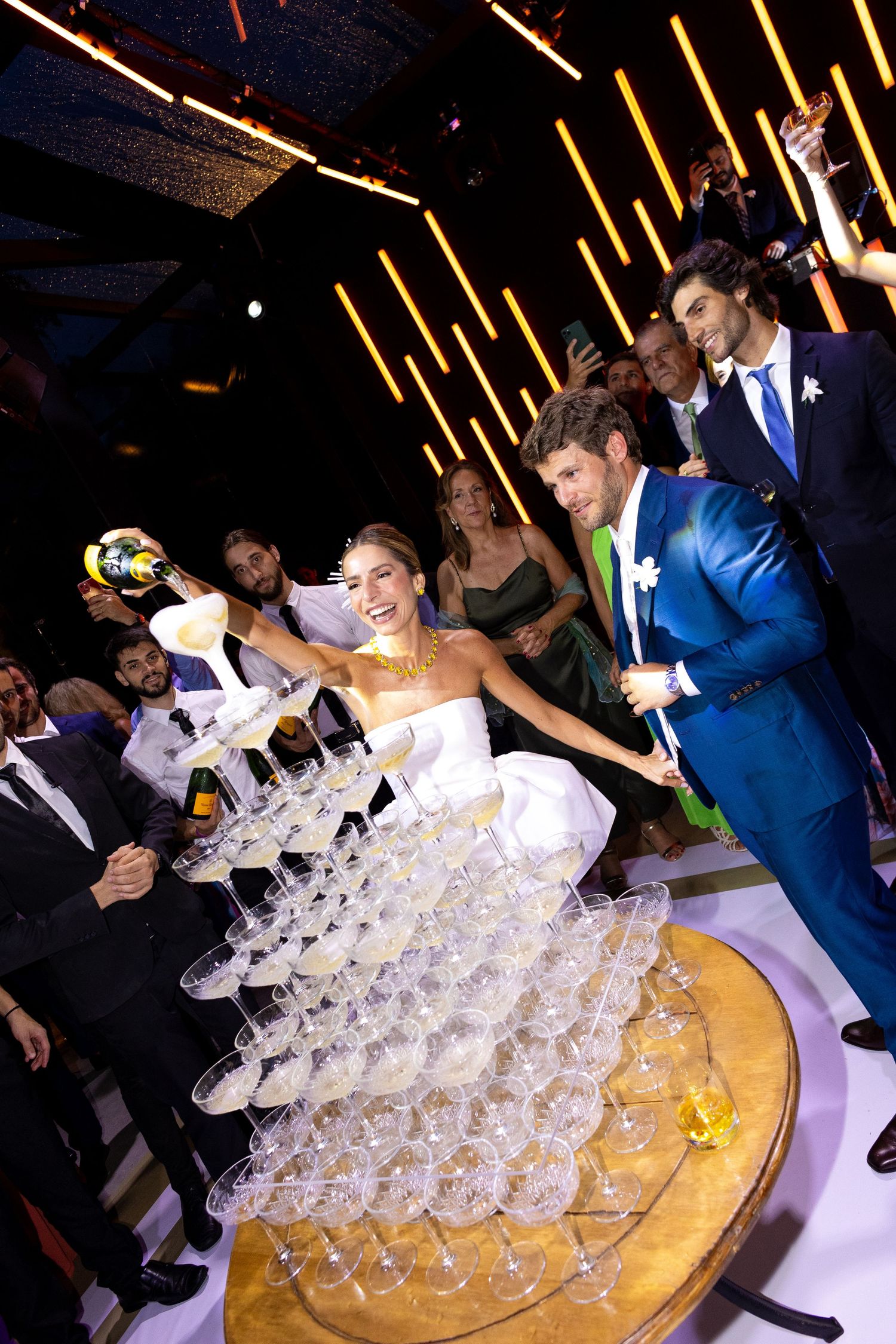 Wedding guests gather around a champagne tower as drinks are poured during an elegant reception celebration.
