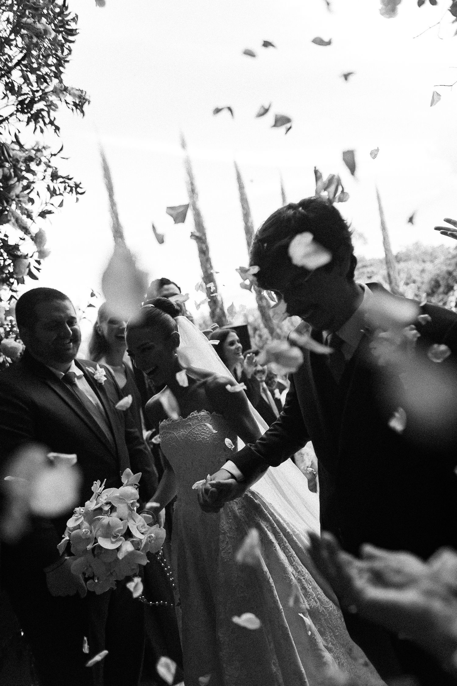 Black and white wedding photo of bride and groom walking through flower petal confetti shower with guests celebrating.