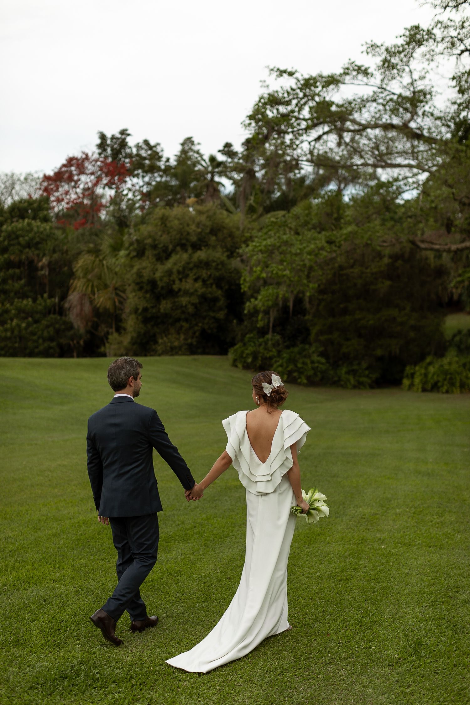 A couple walks hand in hand across a lush green lawn during their wedding.