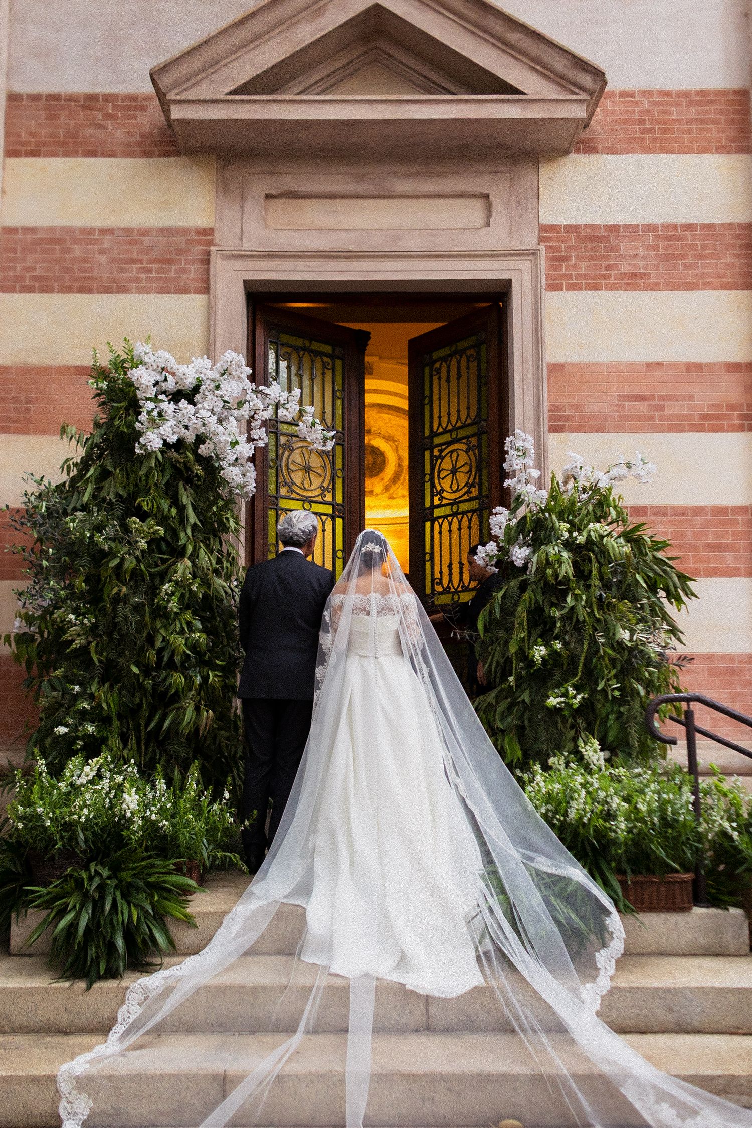 Long flowing white wedding dress veil trails gracefully alongside lush greenery and brick exterior at church entrance.