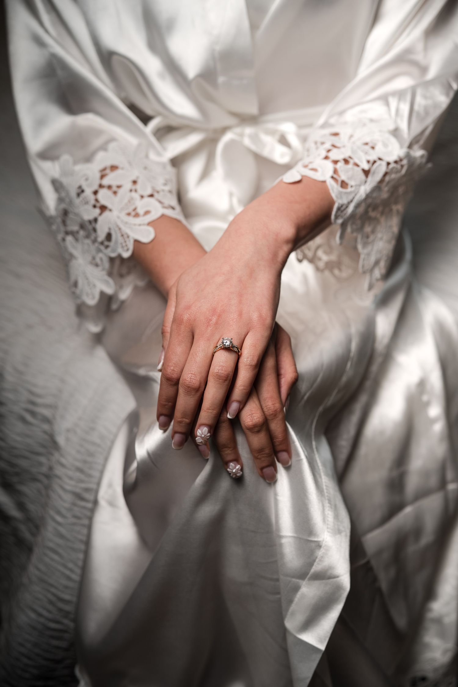Bride in white satin lace robe showing elegant diamond engagement ring on folded hands.