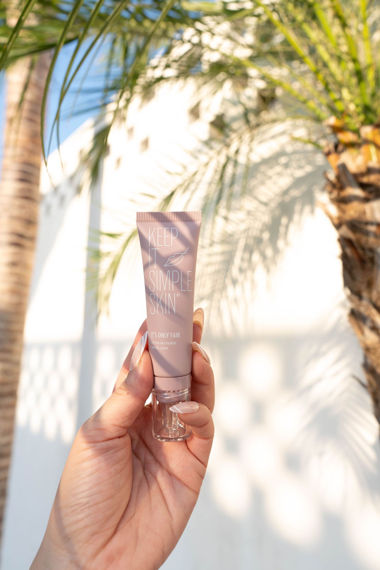 Hand holding up a pink beauty product tube against a white wall with palm tree shadow patterns in sunlight.