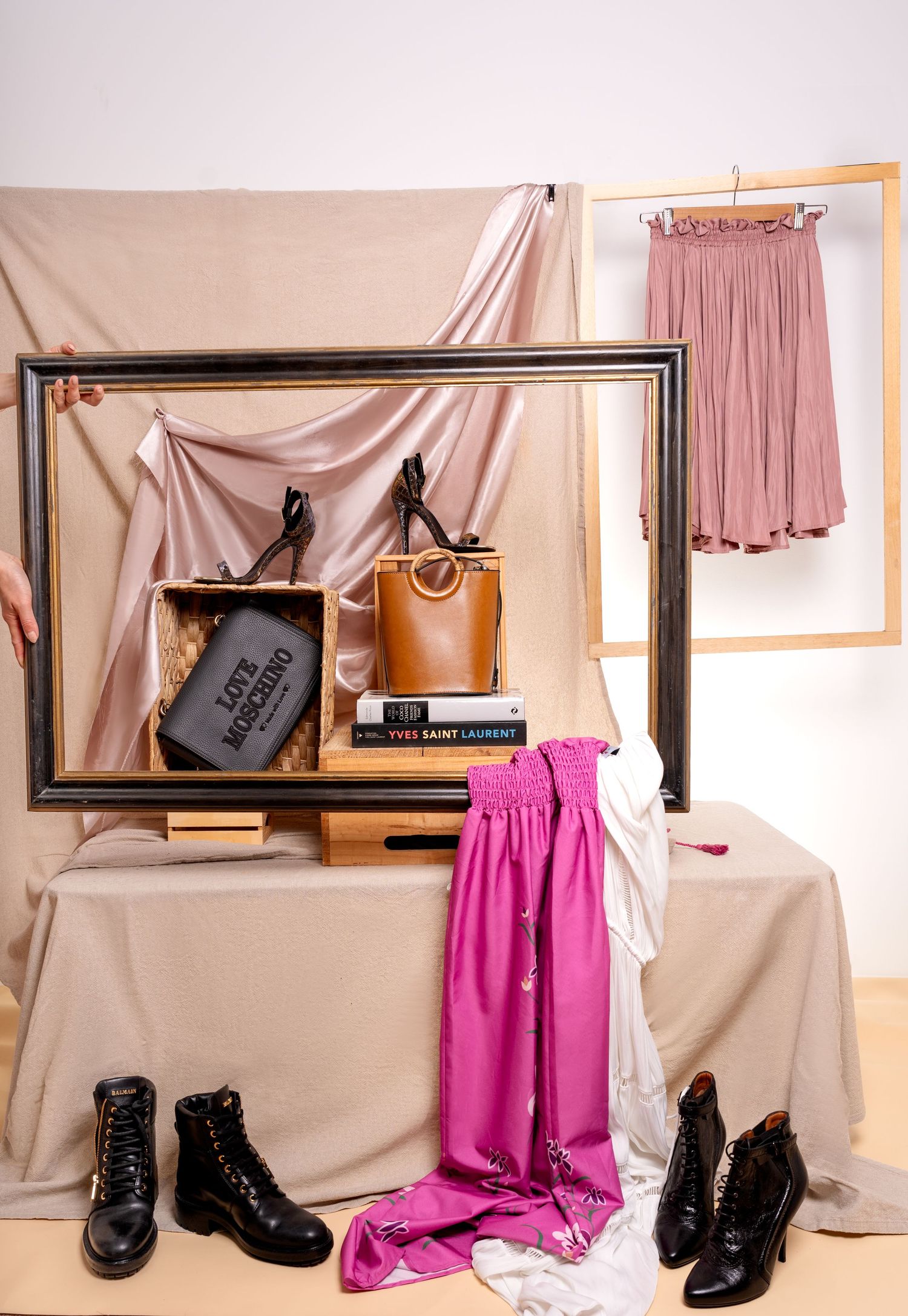 Fashion styling scene with draped blush fabric, black boots, pink skirts, and a brown leather bag against neutral backdrop.