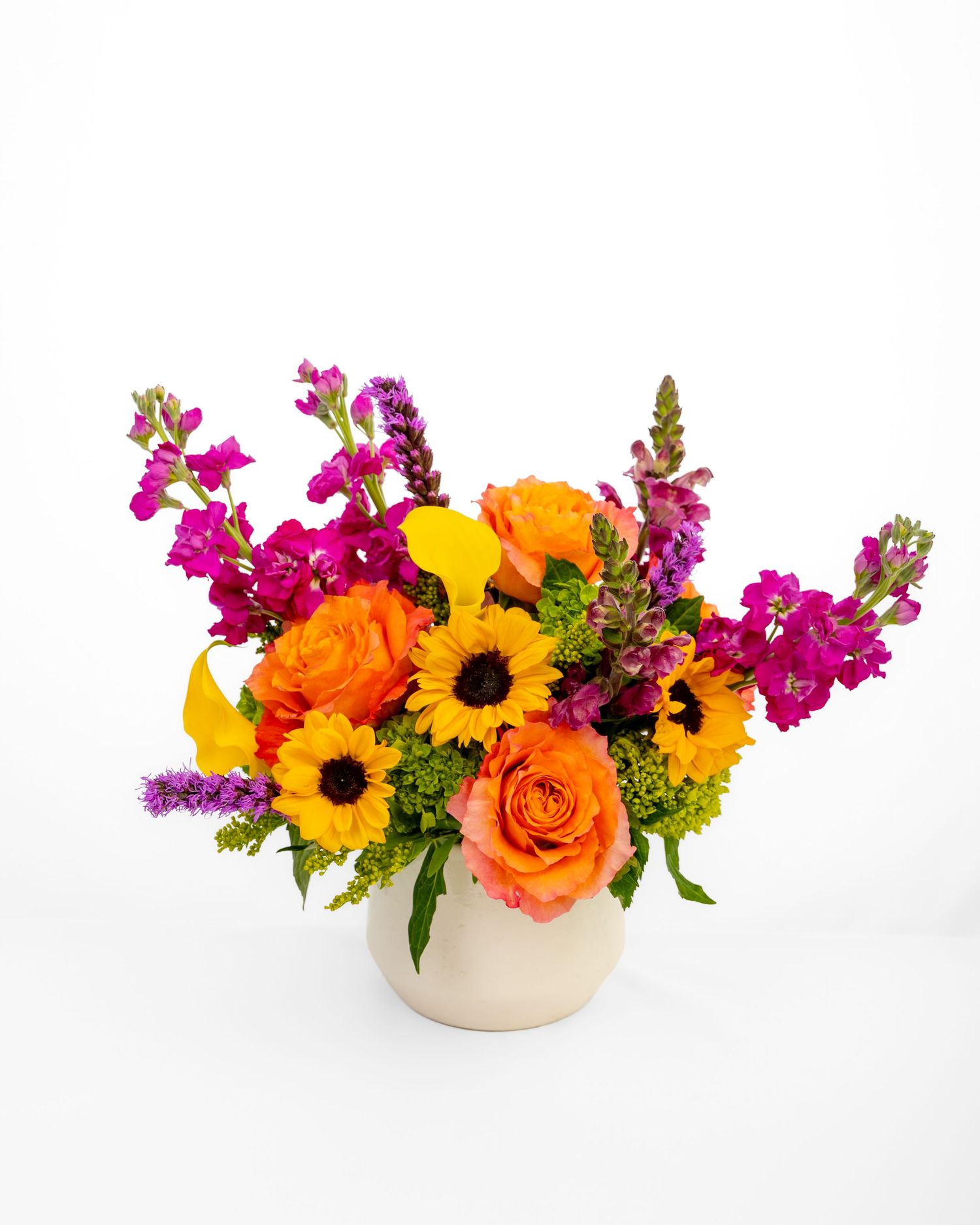 Flower arrangement of oranges, yellows, and purple, including roses, sunflowers, and lilies in a white ceramic vase.