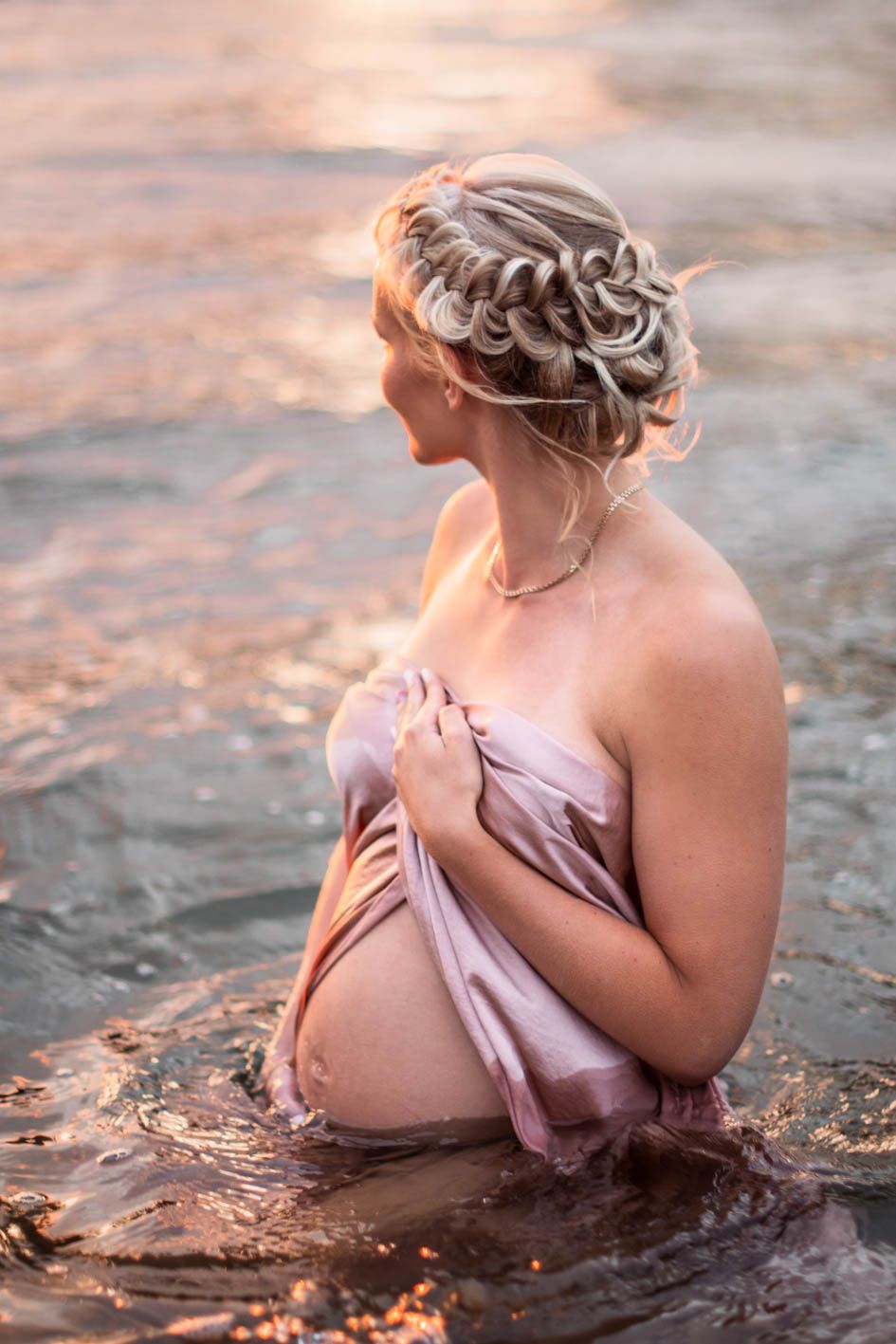 A figure in a pink dress sits gracefully in rippling water during golden hour creating a dreamy artistic portrait.