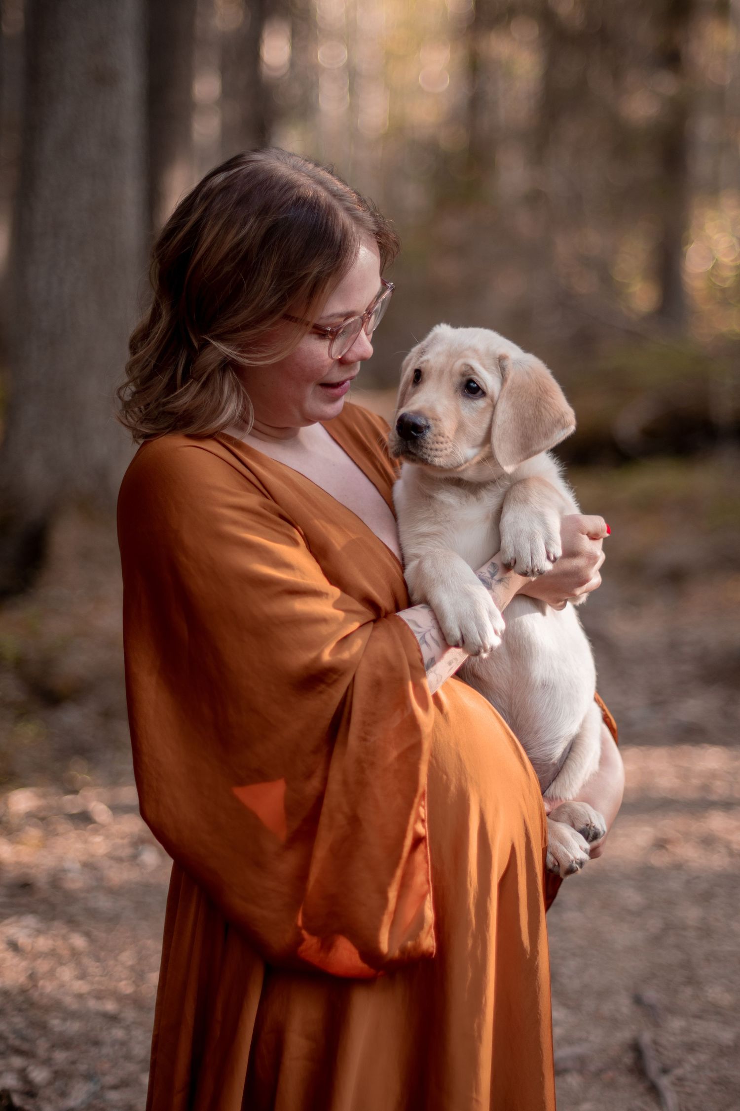 A person in an orange dress holds and embraces a white puppy in a wooded outdoor setting during autumn.