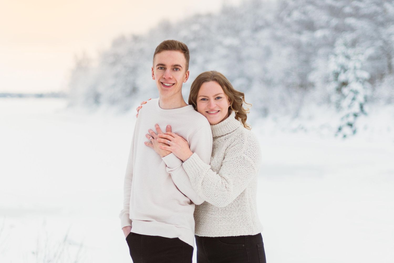 A couple in white sweaters embraces while standing in a snowy winter landscape.