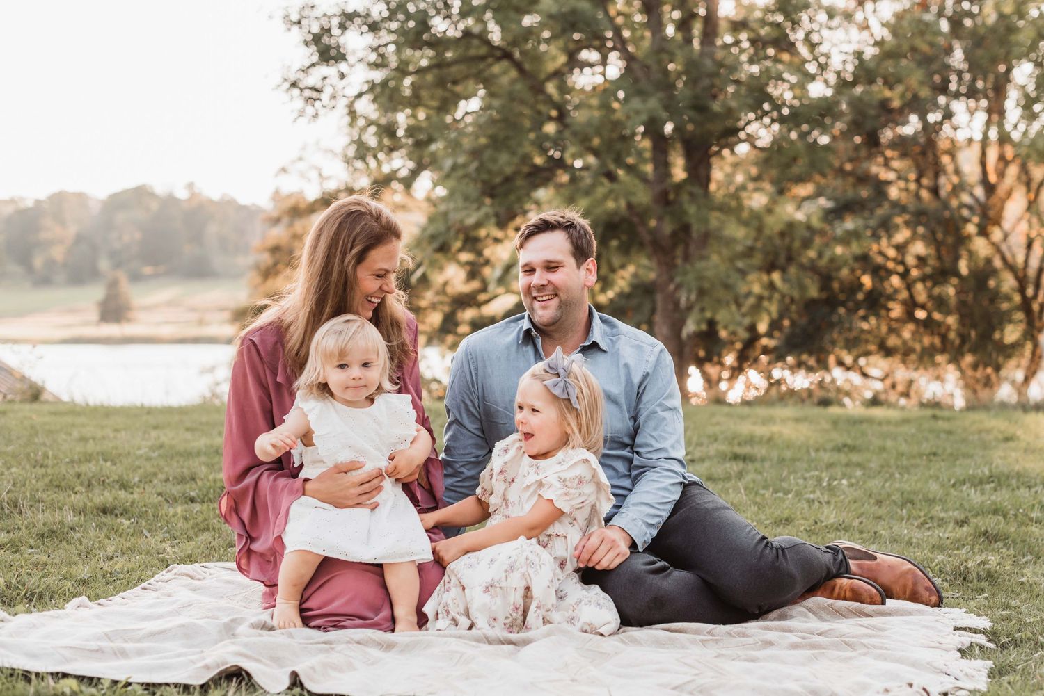 Happy family of four sitting on a blanket outdoors during golden hour with two young daughters.