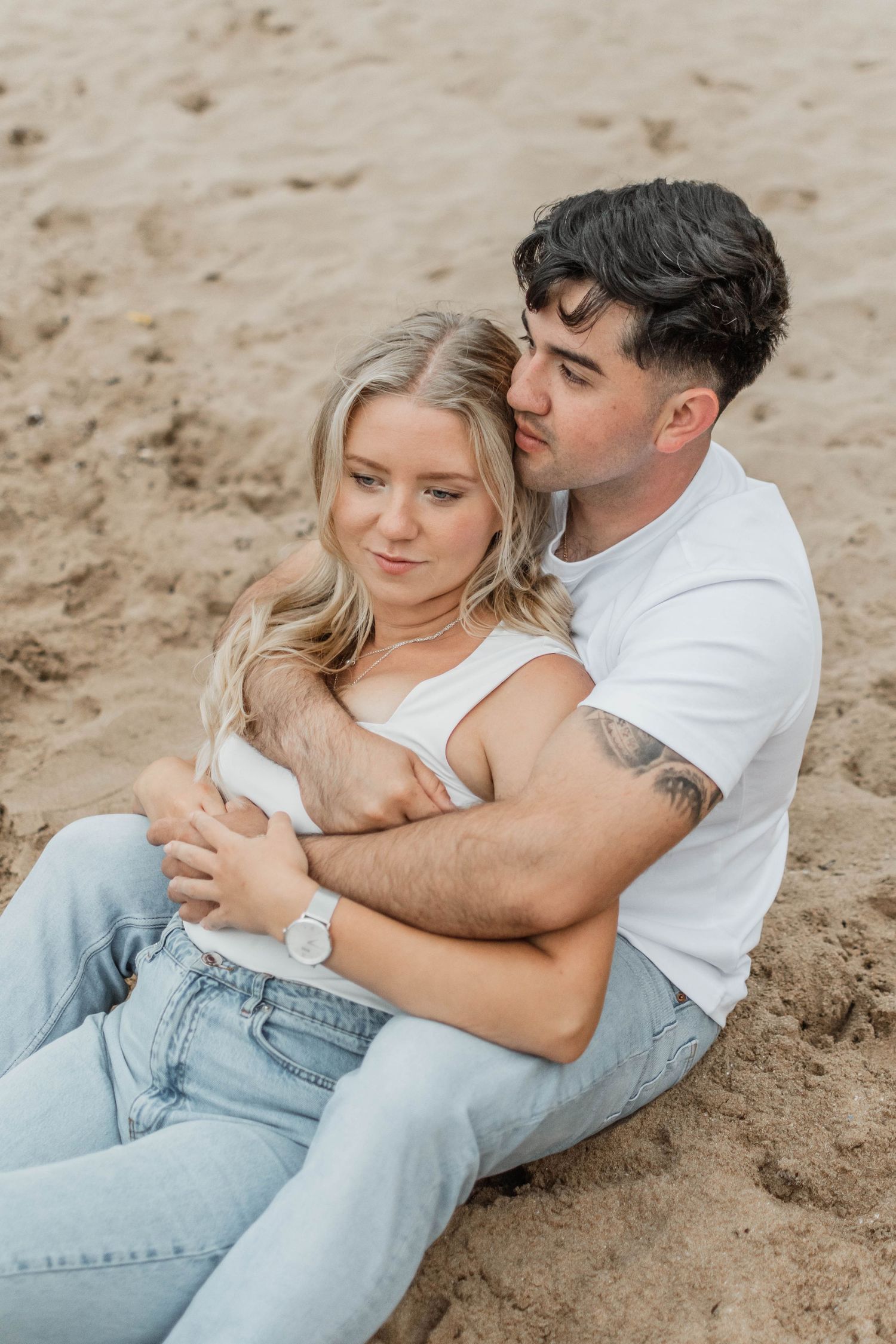 A couple in white shirts and light denim jeans embrace intimately while lying on sandy beach.