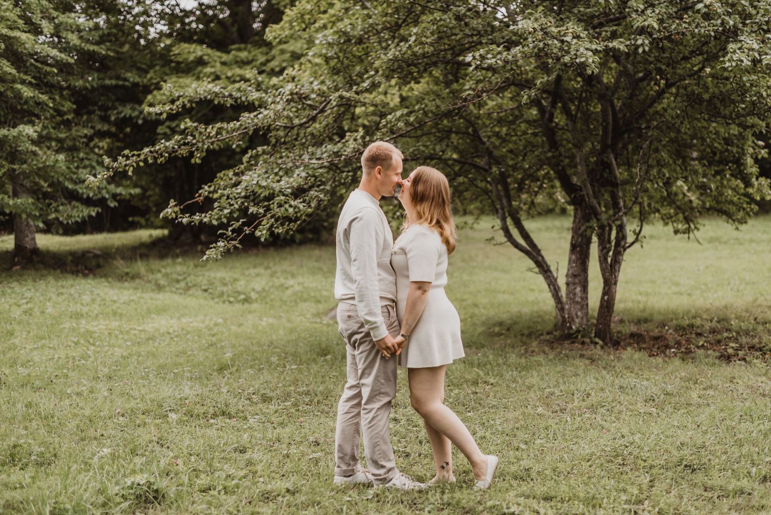 A couple in light-colored attire shares an intimate moment together in a lush green garden setting.