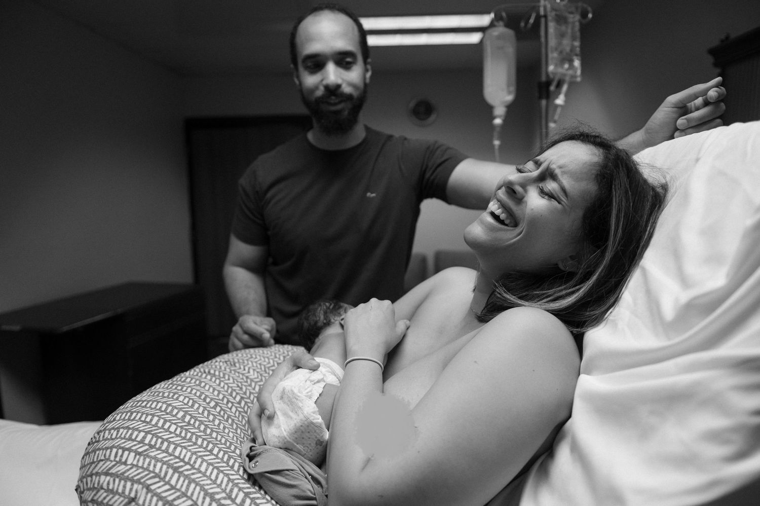 Black and white hospital photo of a joyful new mother holding her newborn while a man stands beside her smiling.