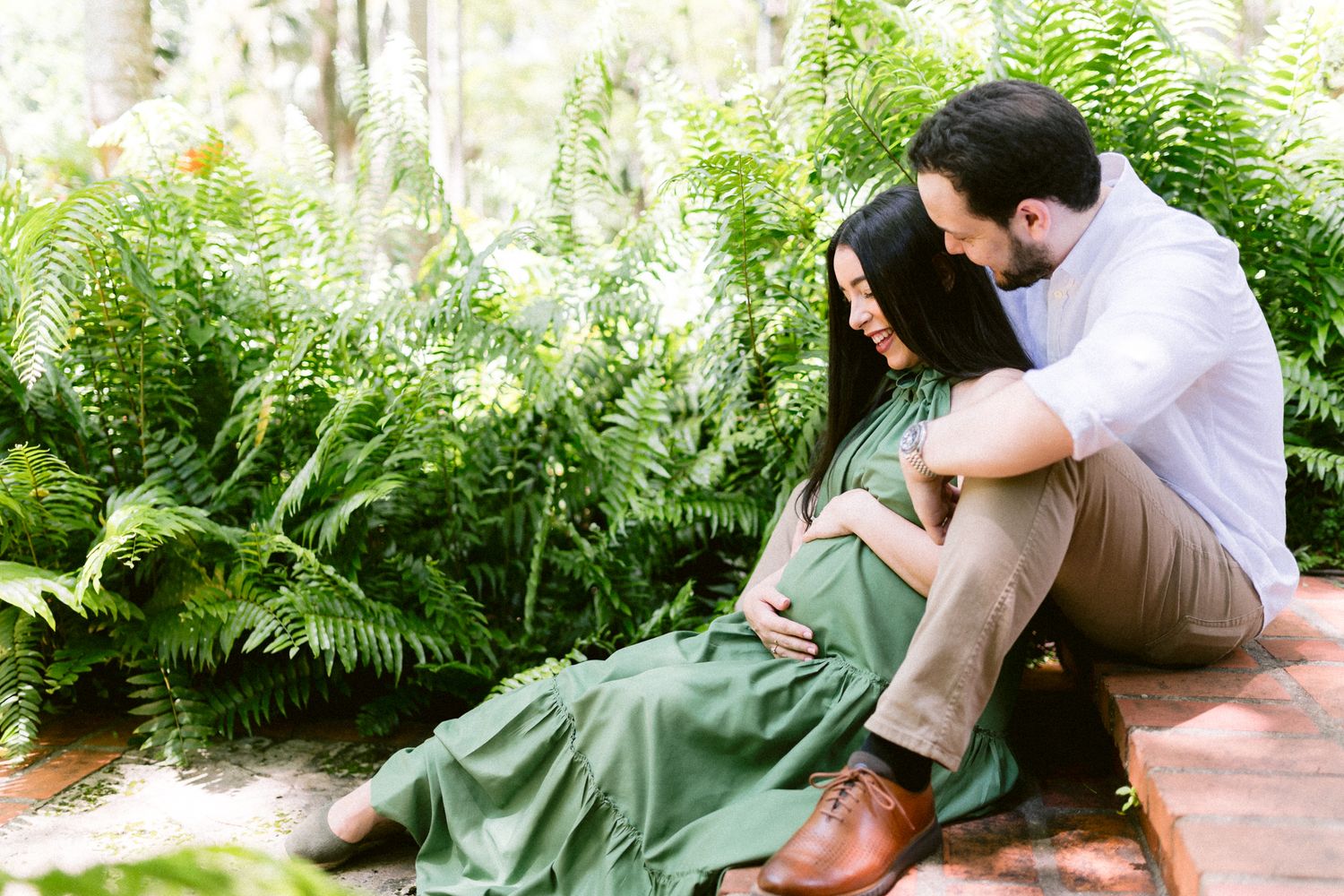 Couple sitting together outdoors among lush green ferns, woman in green dress cradling pregnant belly during maternity photo session.
