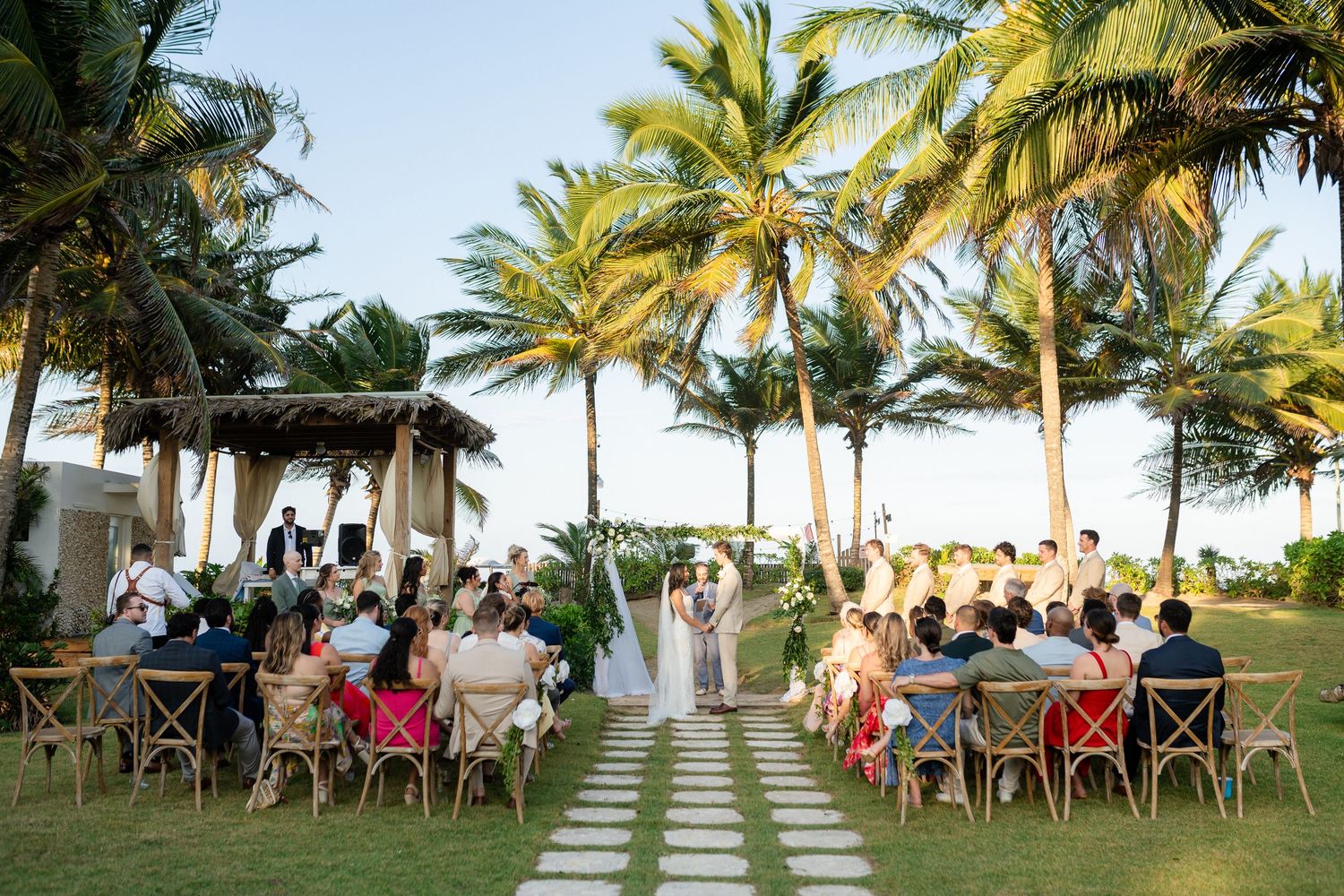 Outdoor beach wedding ceremony with guests seated on wooden chairs along a palm tree-lined aisle at a tropical resort.
