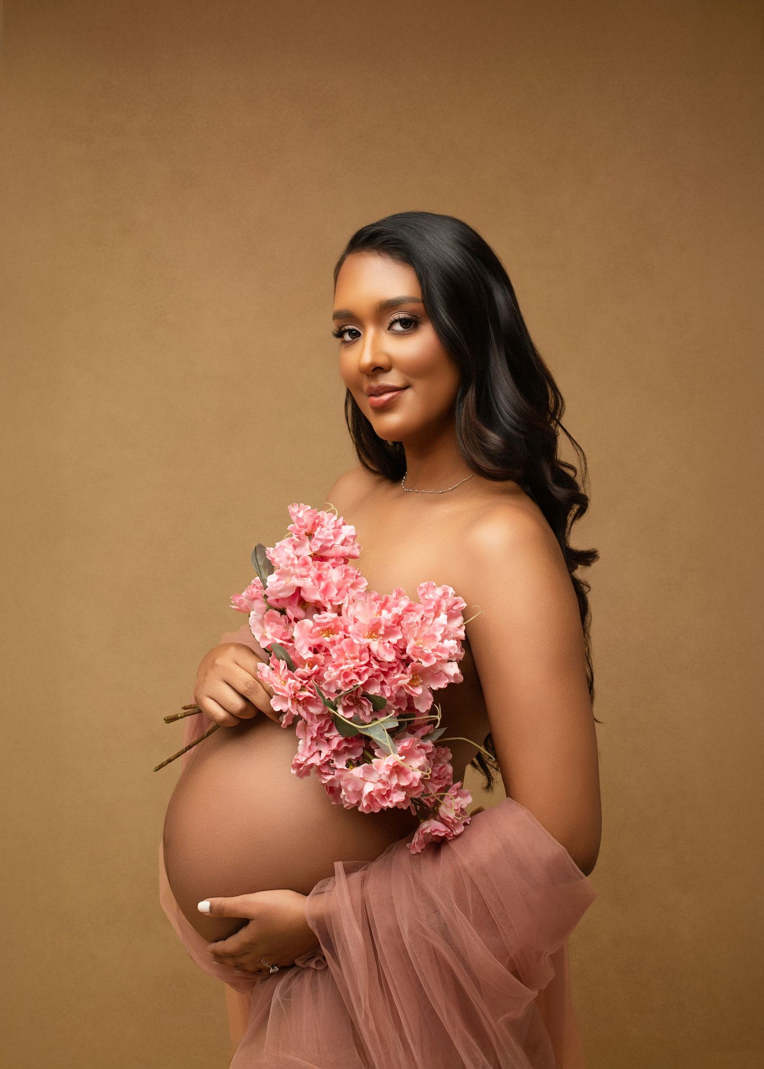A pregnant woman in a flowing pink tulle dress poses with pink flowers against a warm brown backdrop.