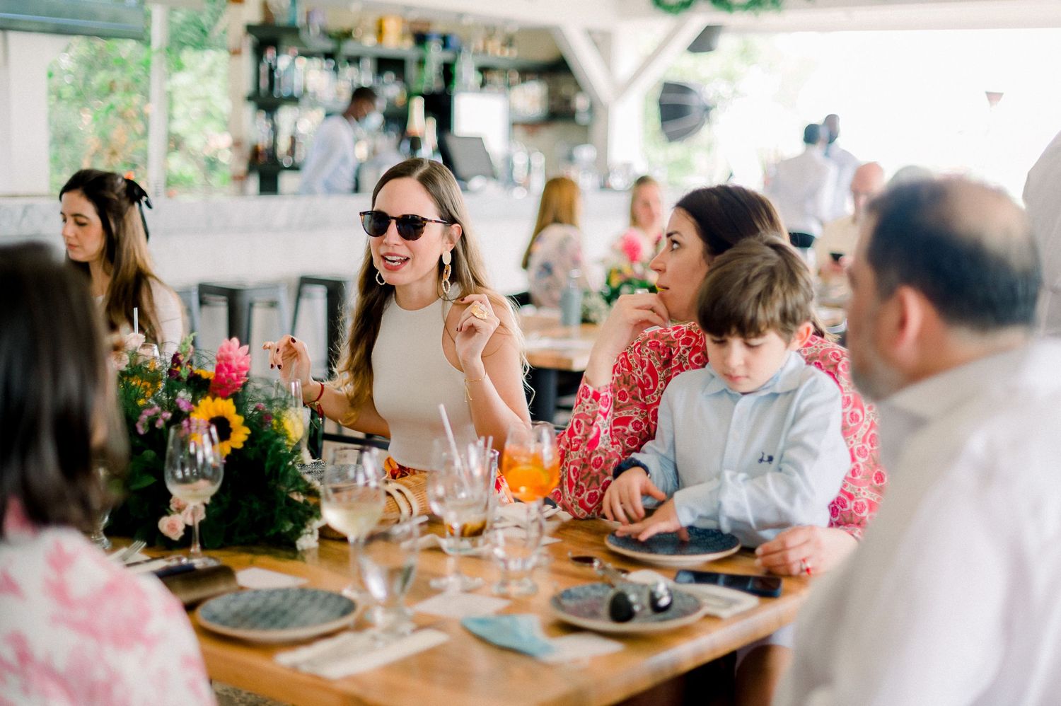 Group of people enjoying a lively outdoor restaurant gathering, smiling woman in sunglasses seated at wooden dining table.