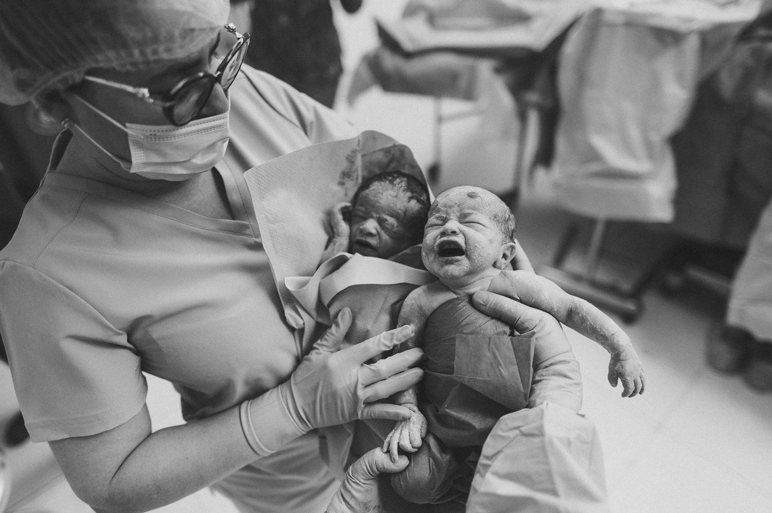 Black and white photo of a nurse holding two crying newborn babies in a hospital delivery room.