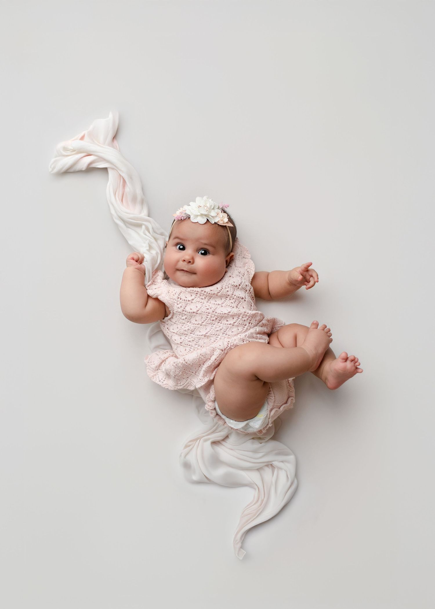 A baby in pink ruffled dress lying on soft white fabric showcases different angles and playful poses during a photo session.