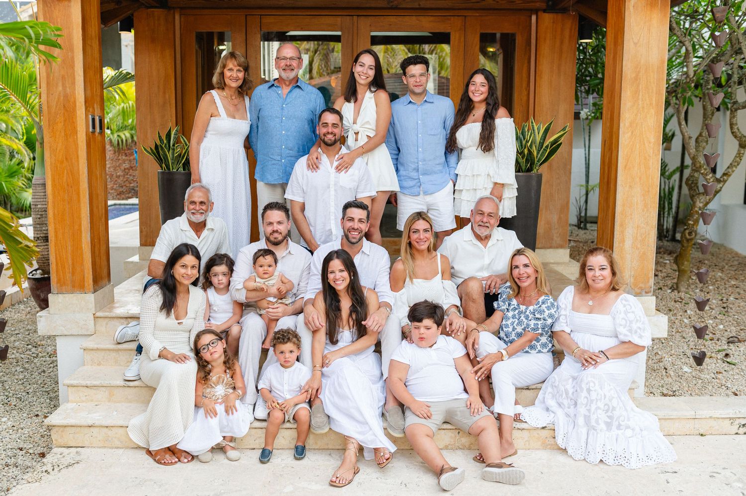 Large family group photo in white and blue outfits posing on steps outside a tropical home.