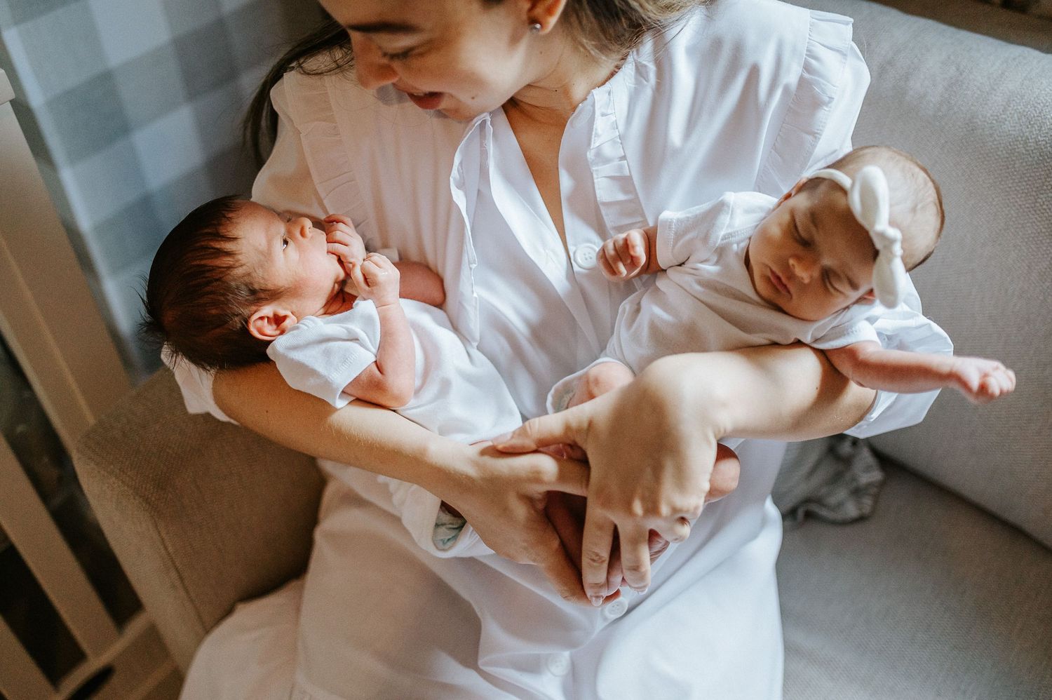 Mother in white dress tenderly holds two sleeping newborn twins in her arms while sitting on a couch.