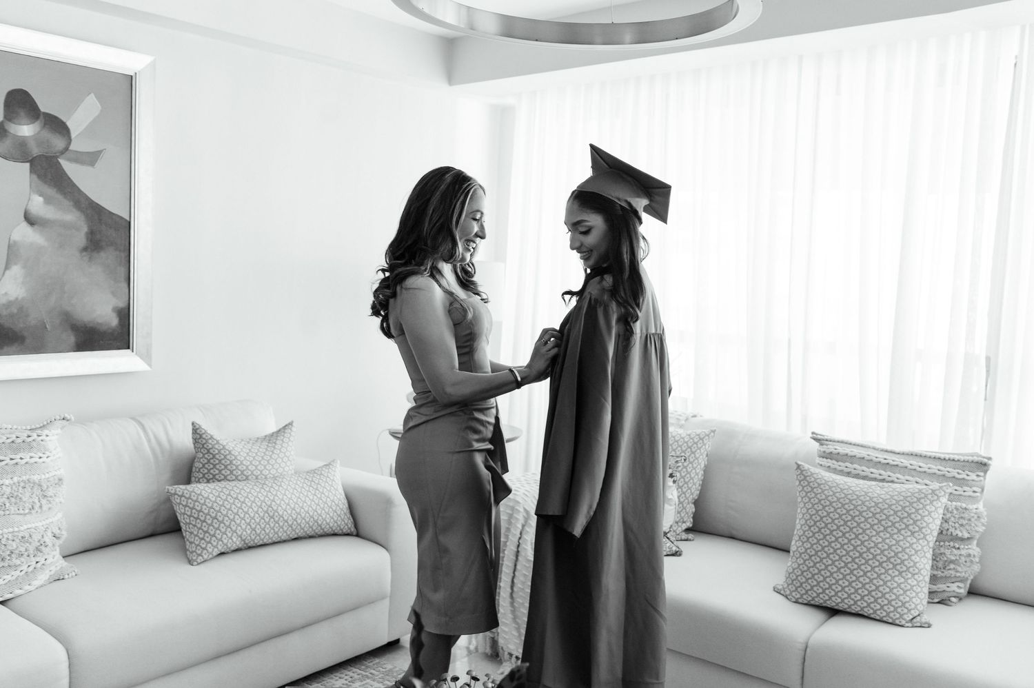A mother helps her daughter adjust graduation gown and cap in a bright living room, captured in black and white.