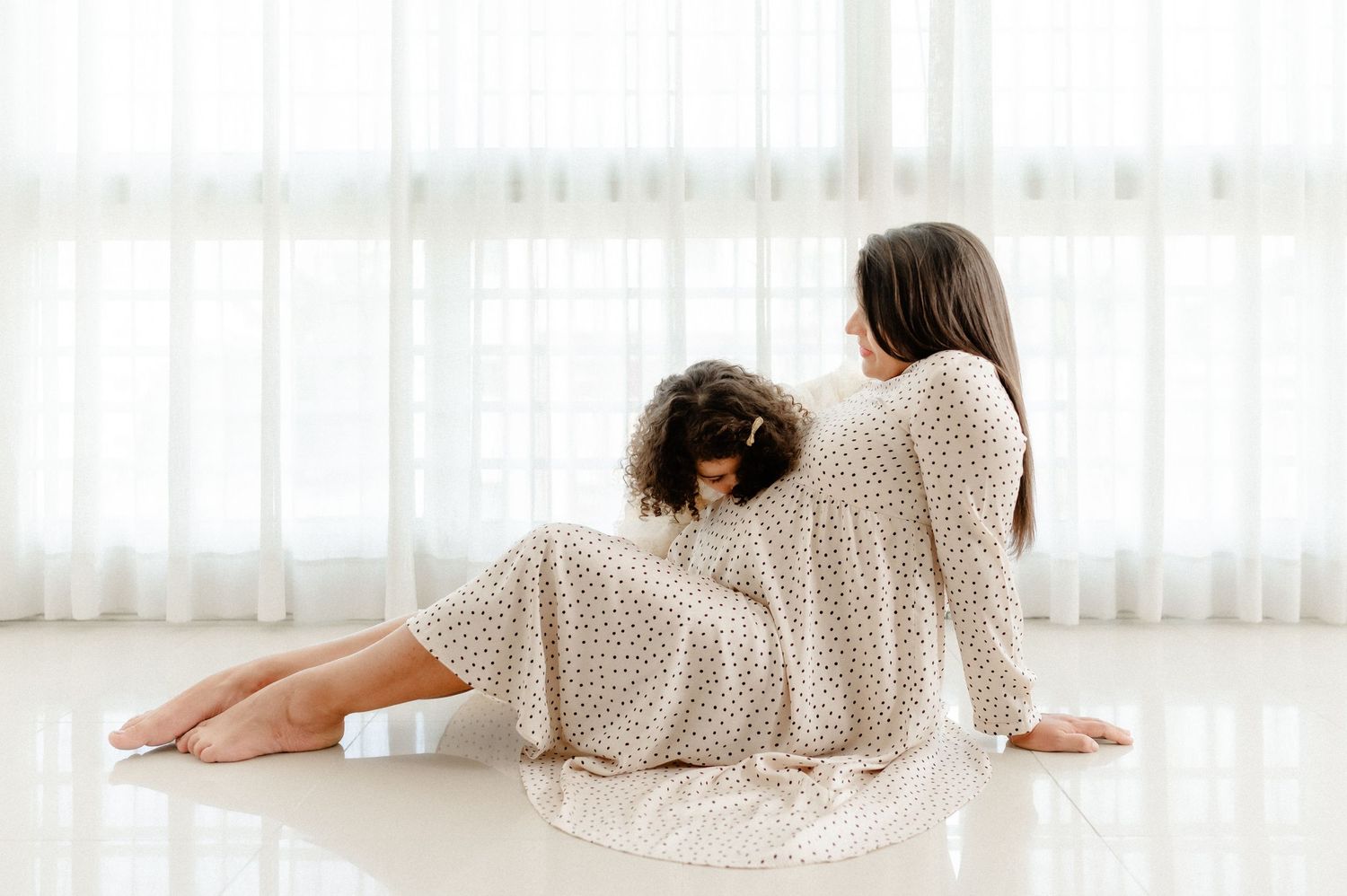 Mother and daughter in matching floral dresses sitting together on floor near white curtains.