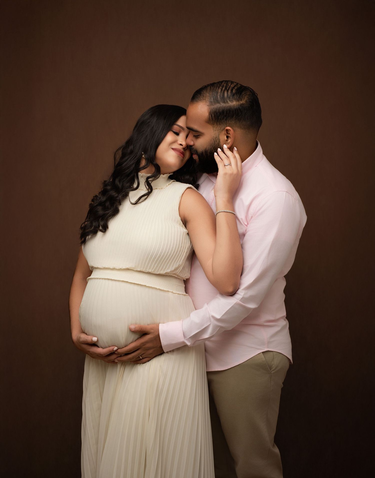 A couple in neutral attire share a romantic moment in matching cream and beige tones against a dark brown studio backdrop.