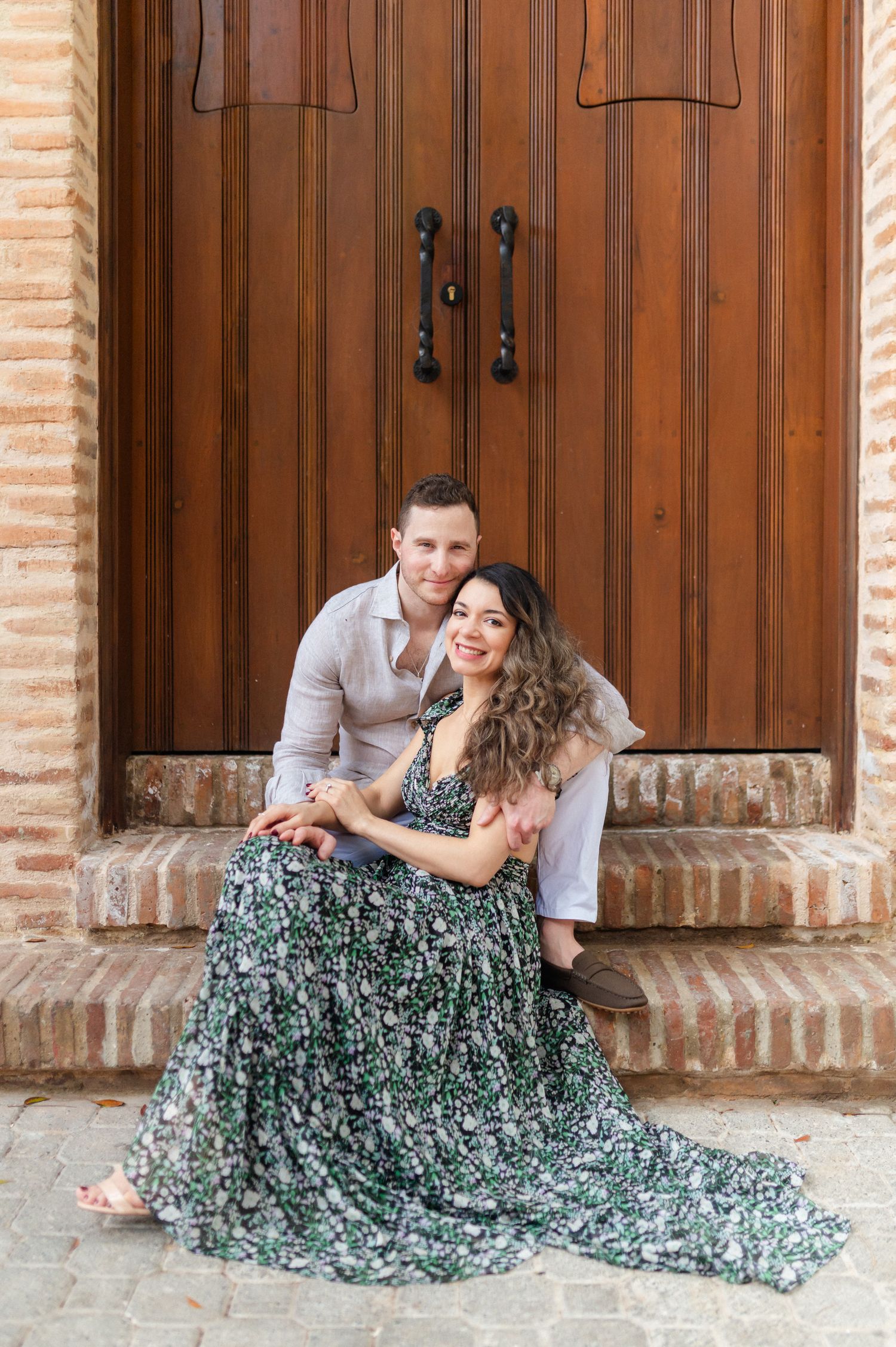 Couple sitting on brick steps in front of large wooden doors, woman in floral green dress, man in light gray shirt.