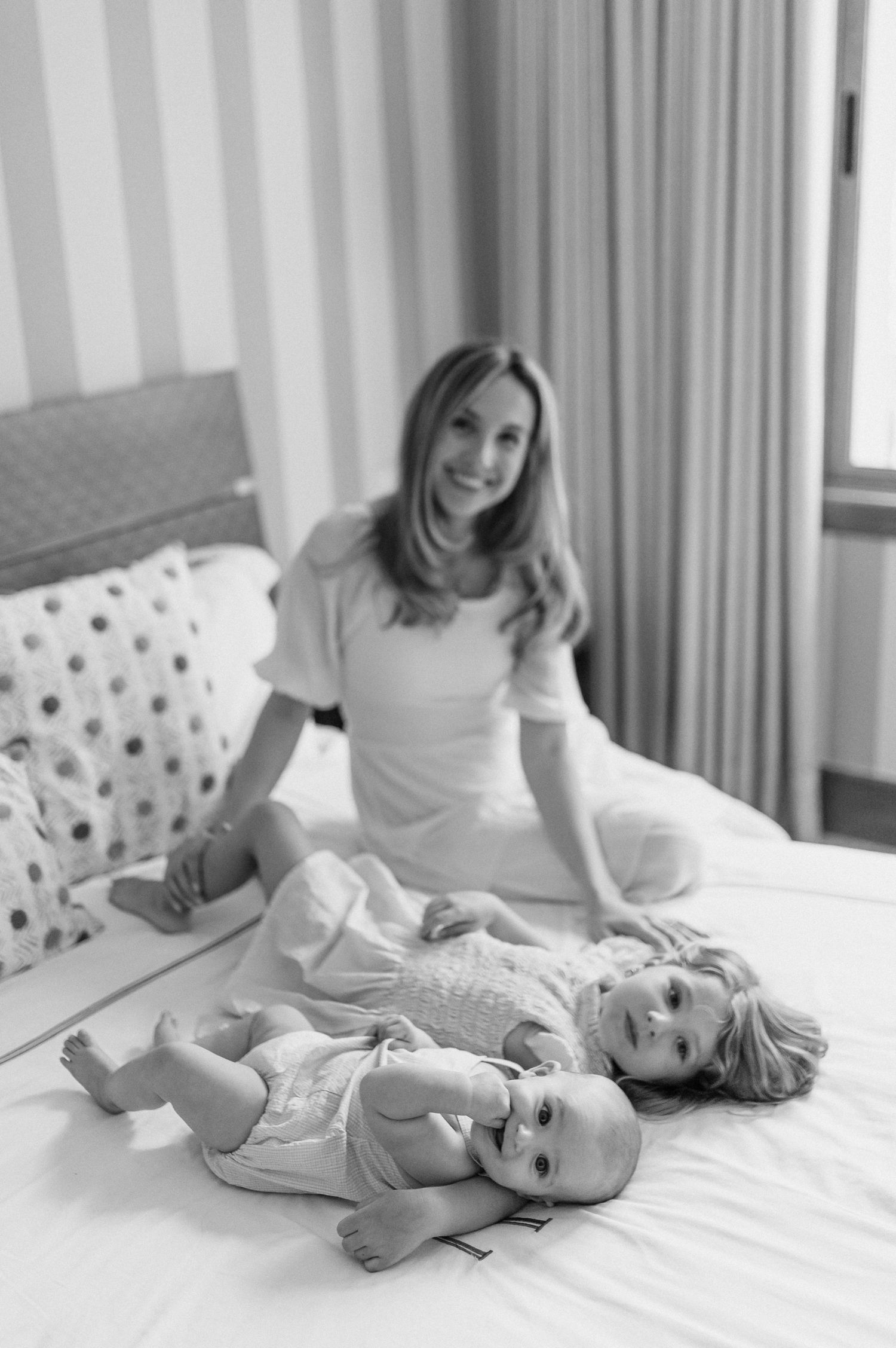Black and white photo of a smiling mother sitting on a bed with two young children, one baby and one toddler.