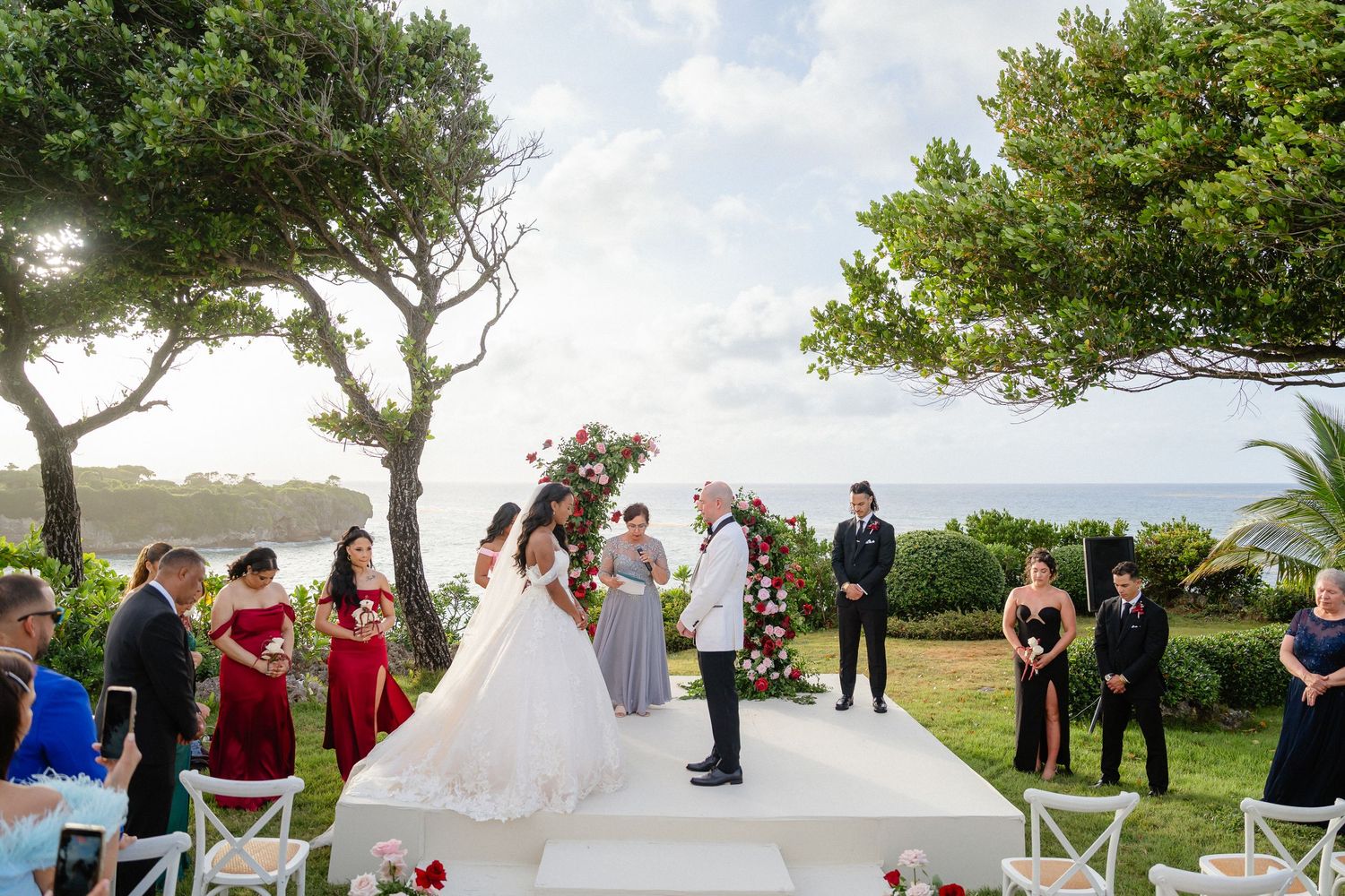 Outdoor wedding ceremony by the sea with bride and groom at floral arch, guests in formal attire on white aisle.
