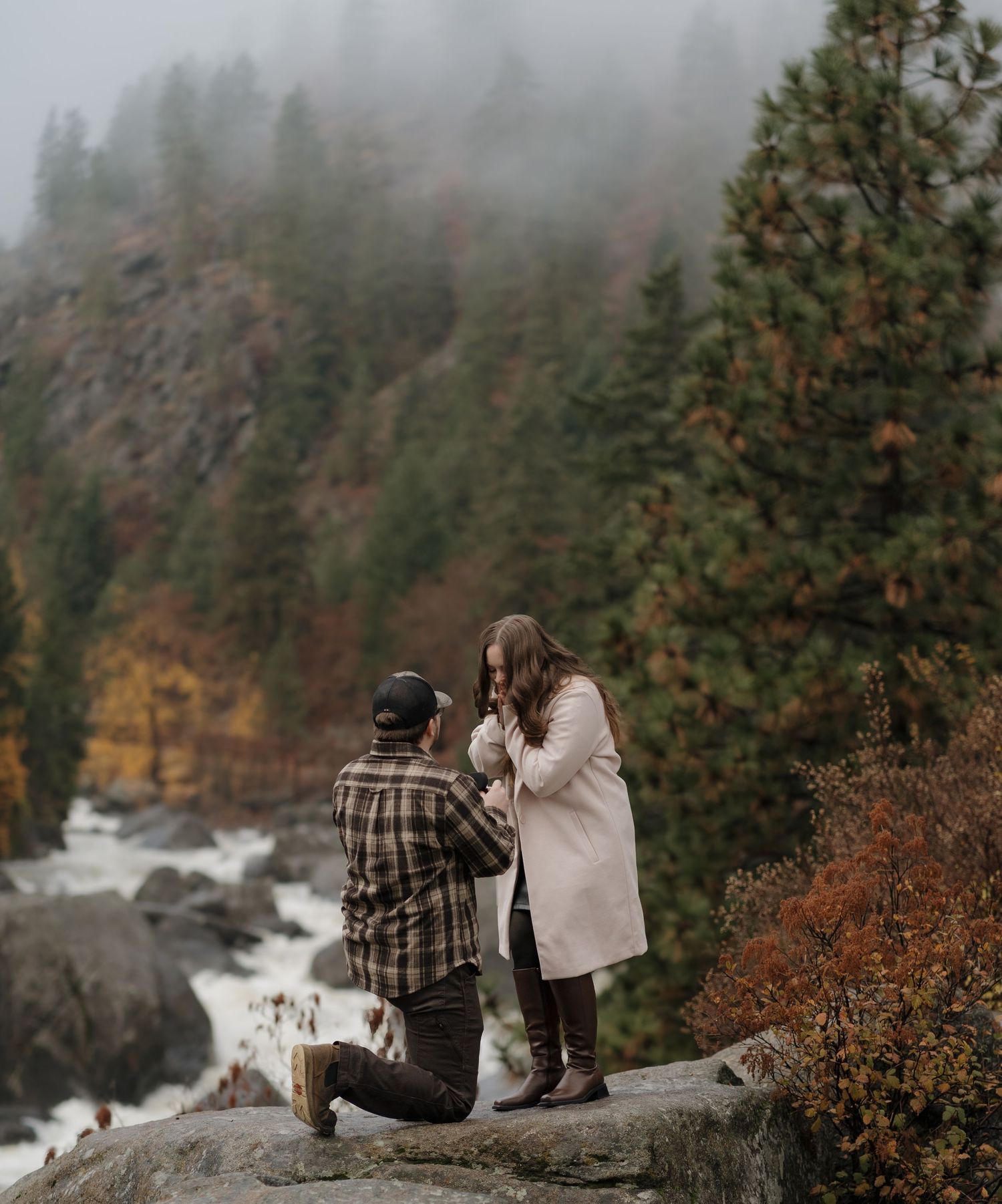 Man kneeling proposing to woman on rocky overlook with misty forest and river backdrop in autumn.
