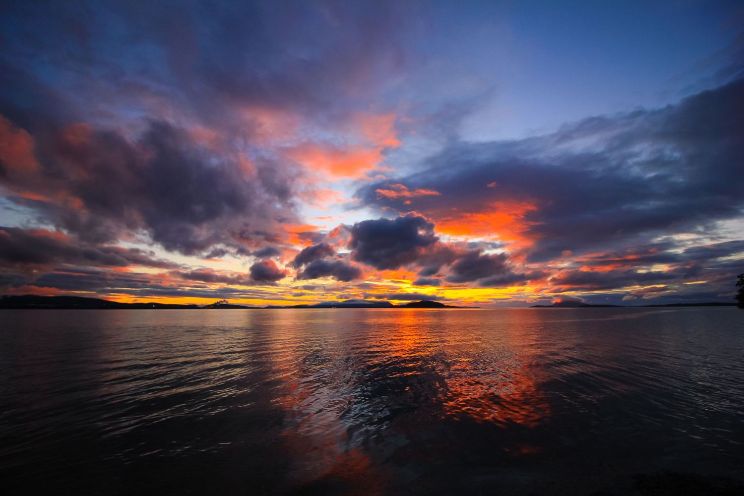 Dramatic sunset over a lake with vibrant orange clouds reflecting in rippling water.
