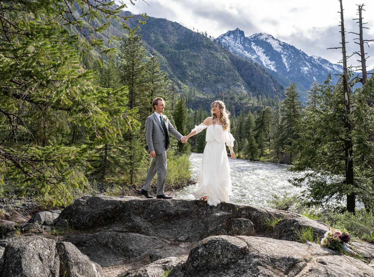Newlyweds standing on rocky terrain with snow-capped mountains and forest in background.