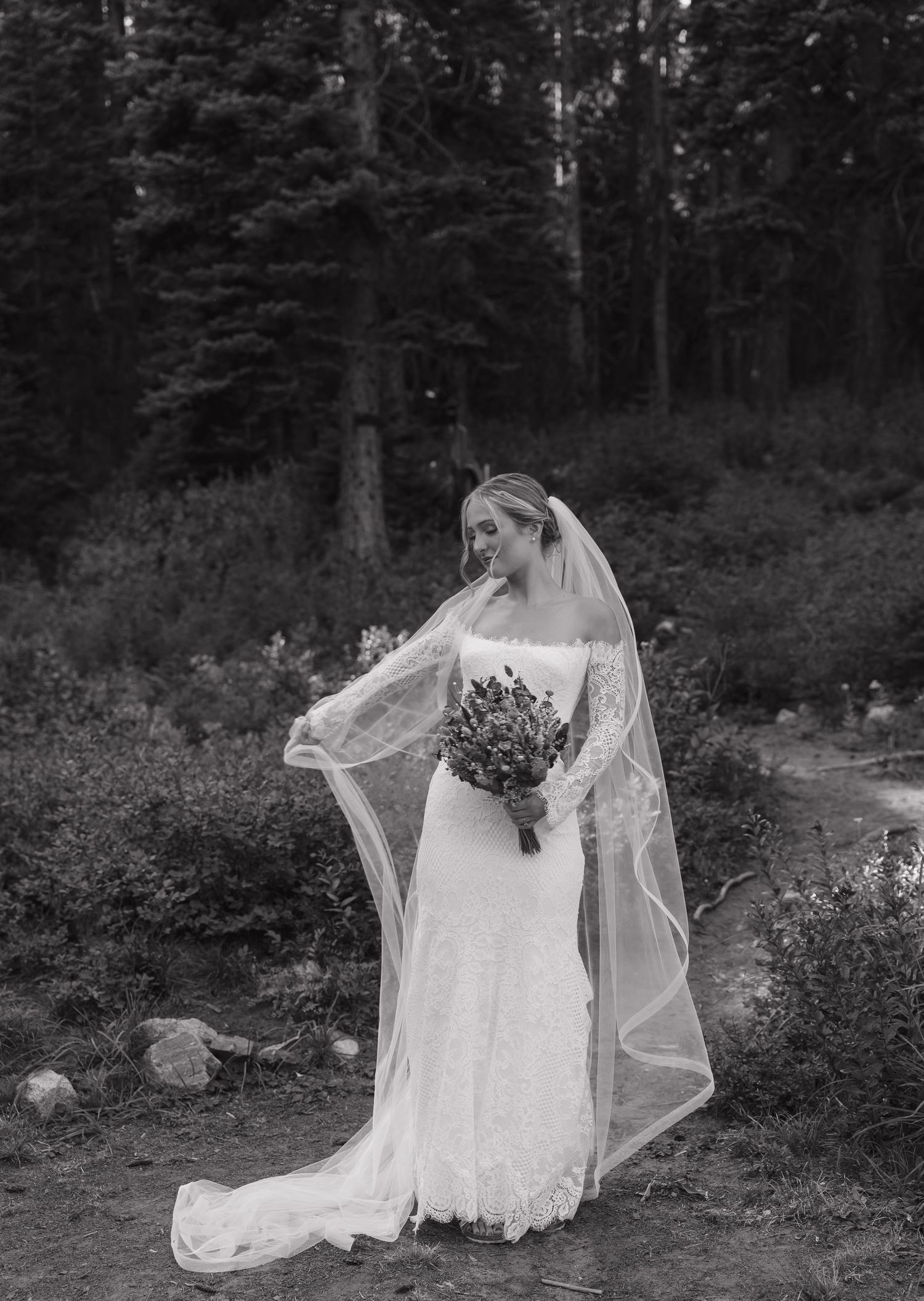 A bride in a flowing white lace gown and veil poses elegantly in a wooded garden setting at dusk.
