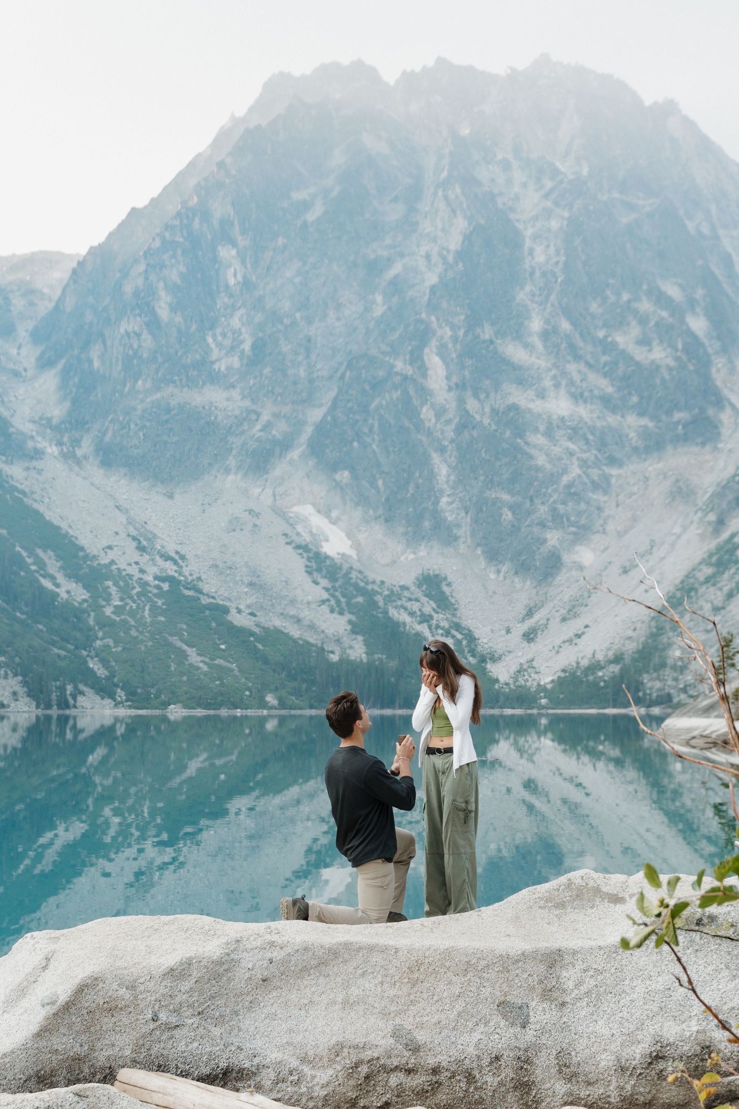 Man proposes on one knee to woman on rocky lakeside with stunning mountain reflection backdrop.