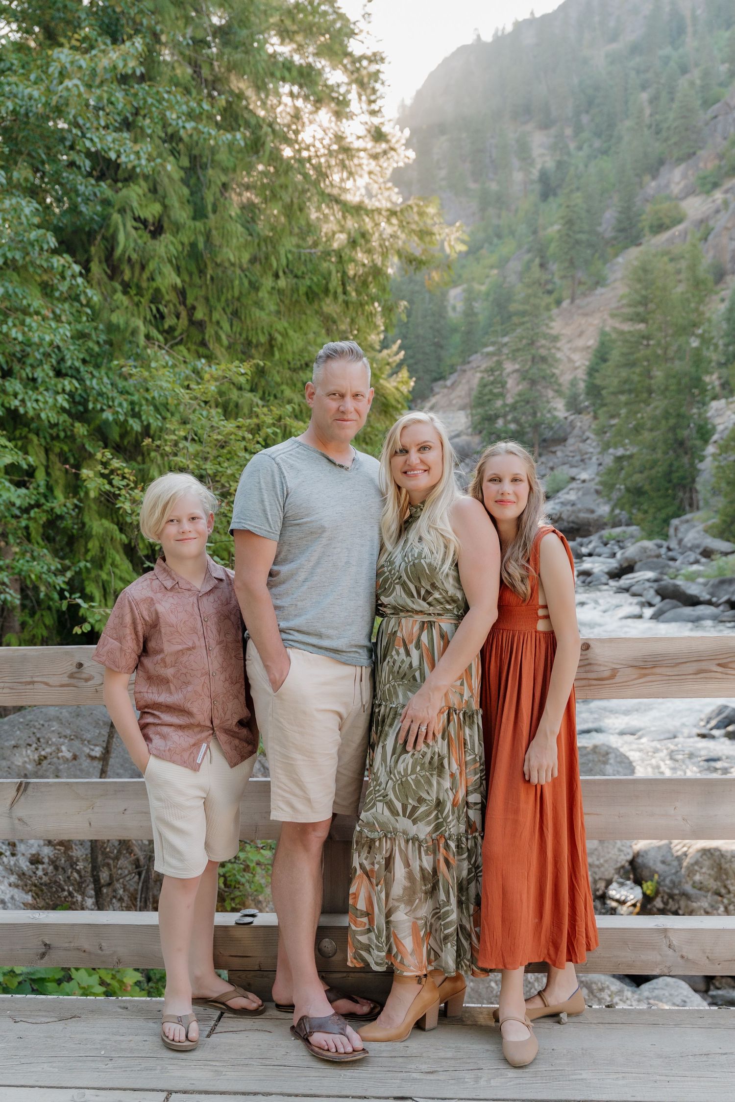 Family of four posing on a wooden bridge with a mountain river and evergreen forest backdrop.