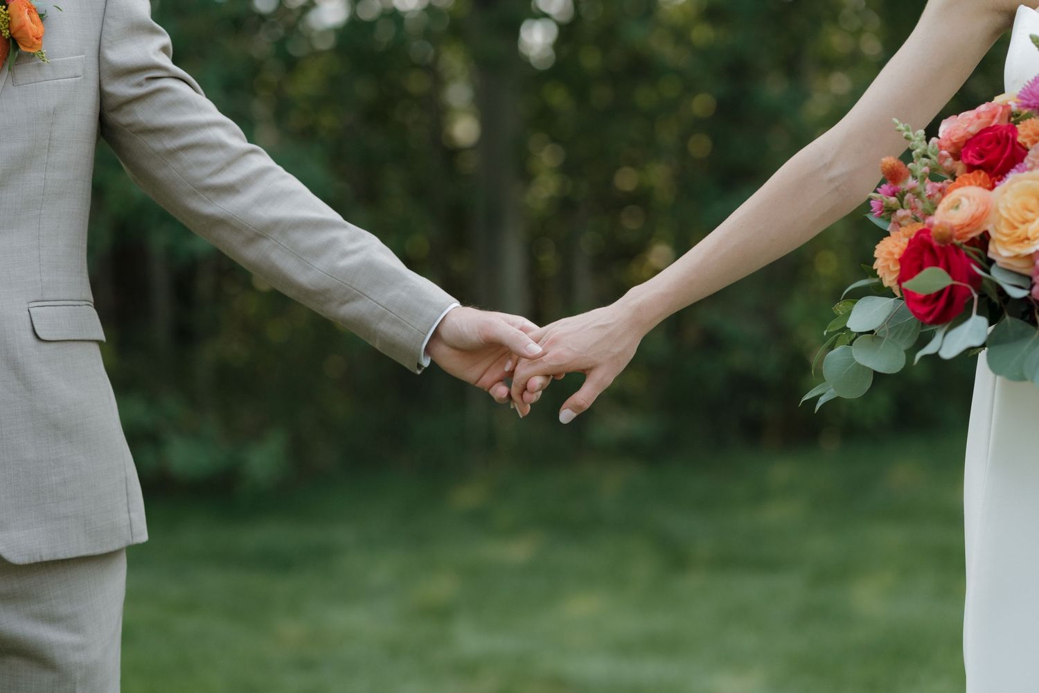 A couple holds hands in a romantic outdoor garden setting with coral and pink floral bouquets.