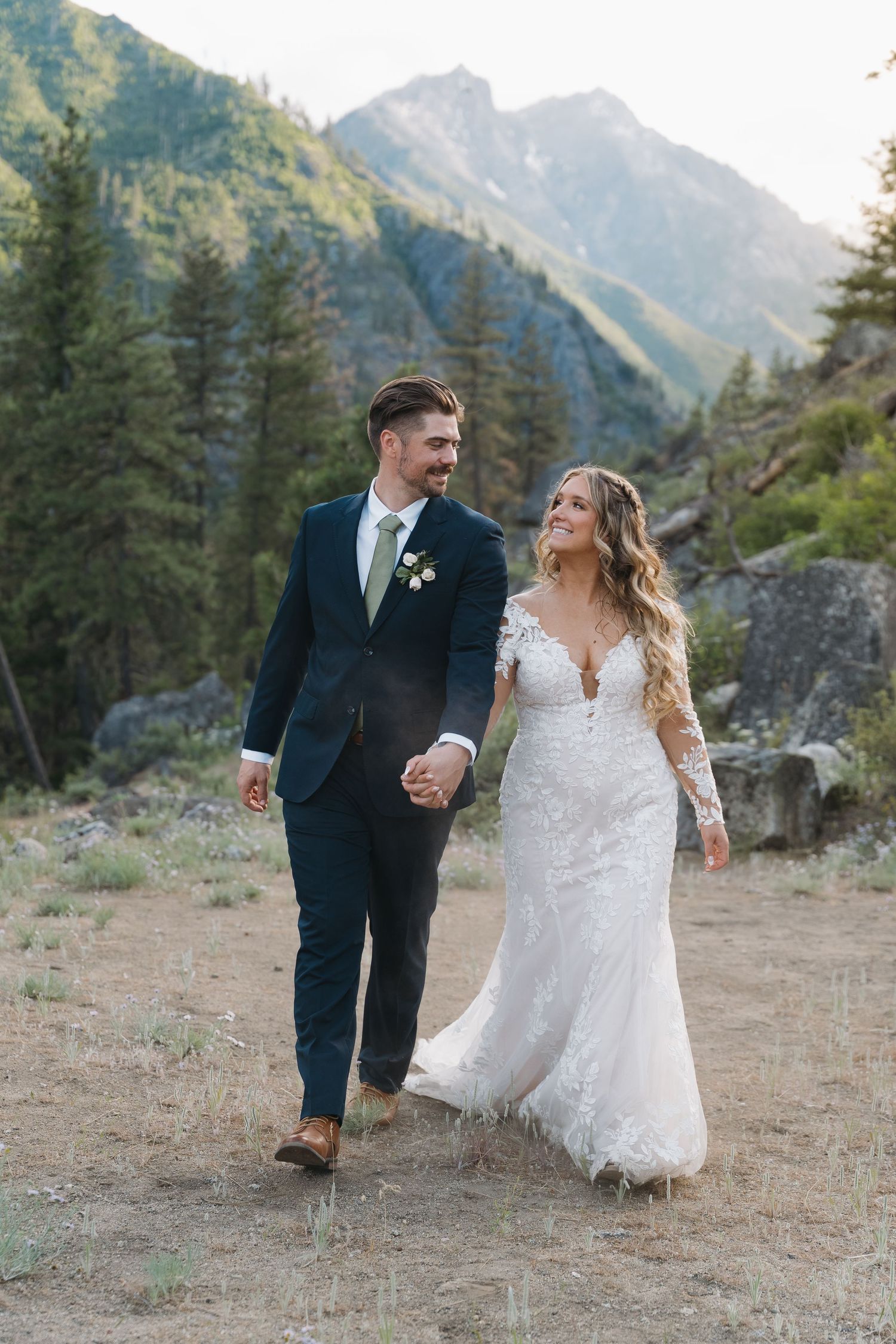 A couple in wedding attire walks hand in hand through a scenic mountain landscape during their outdoor celebration.