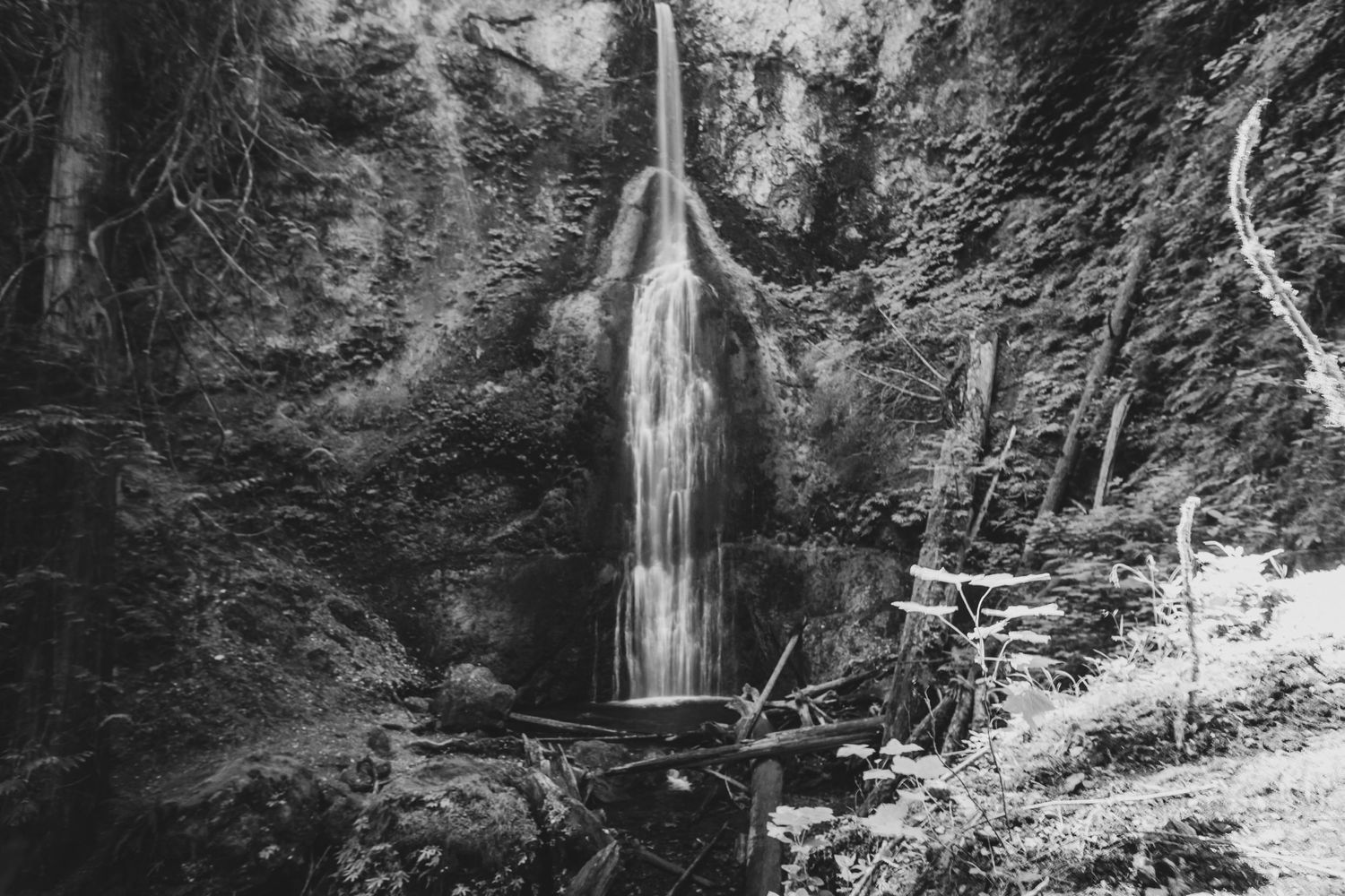 Black and white photograph of a tall waterfall cascading between rocky cliffs with a wooden walkway.