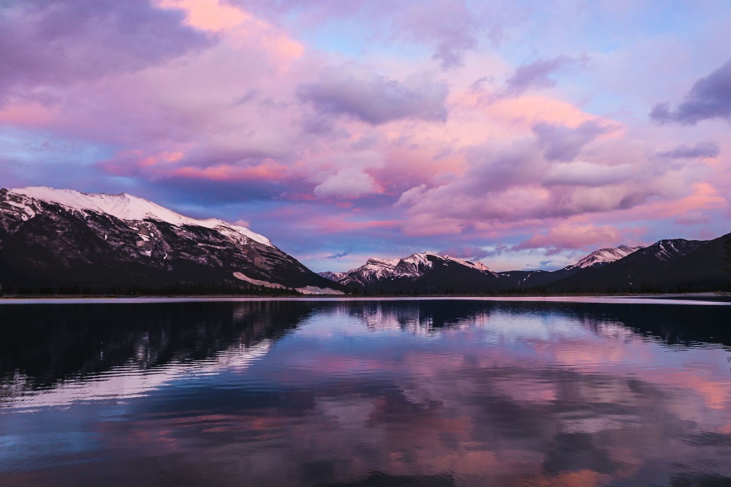 Snow-capped mountains reflect in lake waters under dramatic pink and purple sunset clouds.