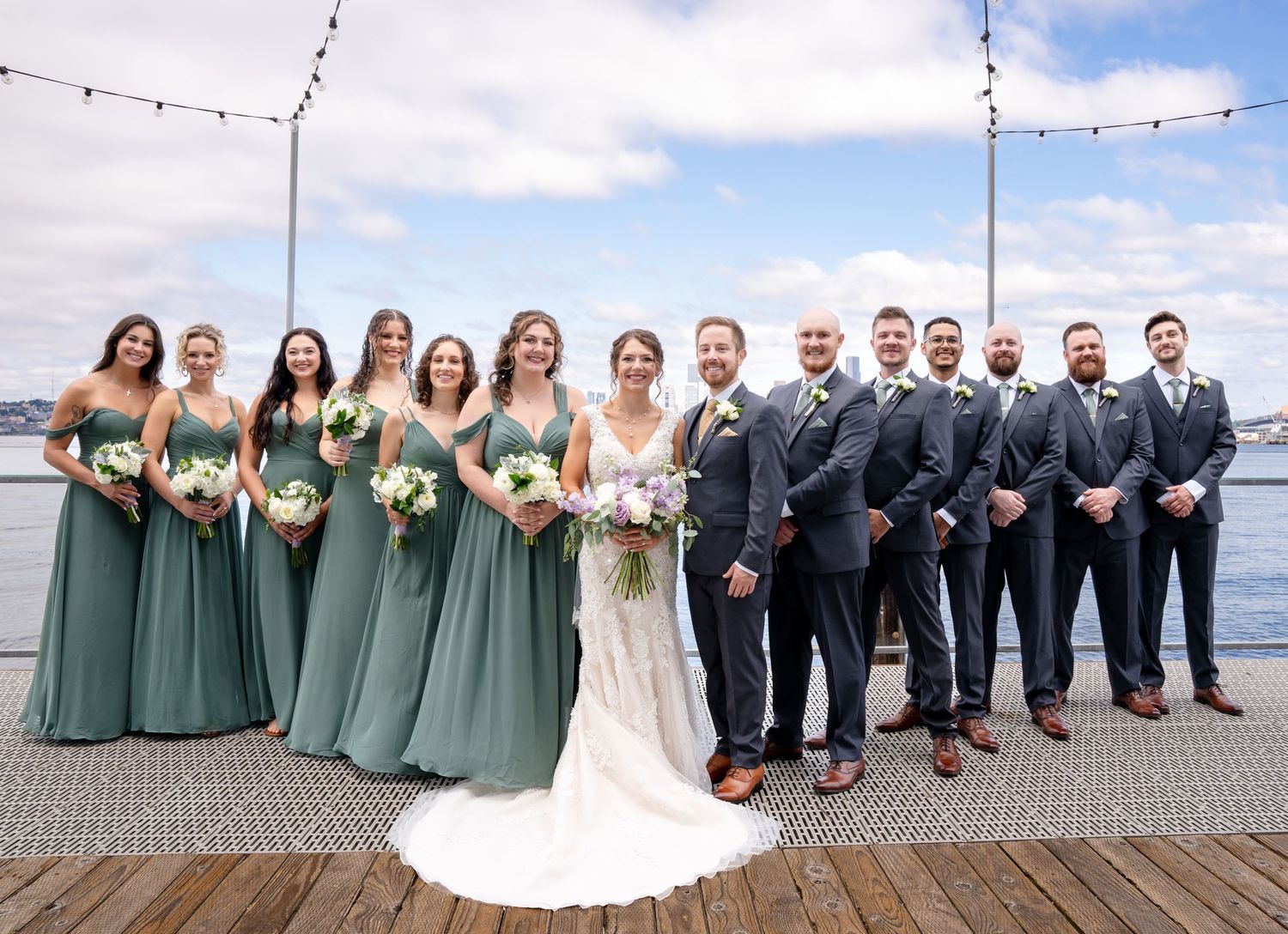 Wedding party in sage green dresses and navy suits posed on waterfront deck.