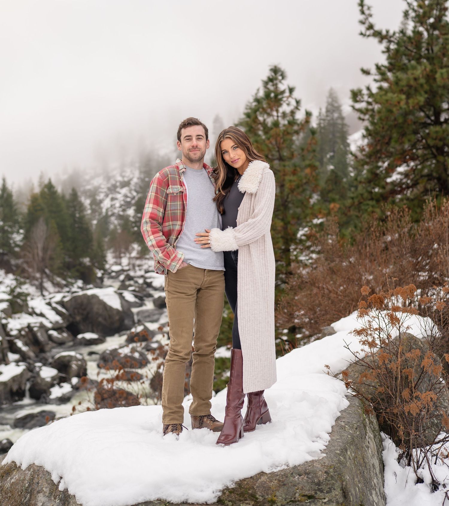 A couple poses together in a snowy mountain landscape with evergreen trees and boulders in the background. Leavenworth, WA